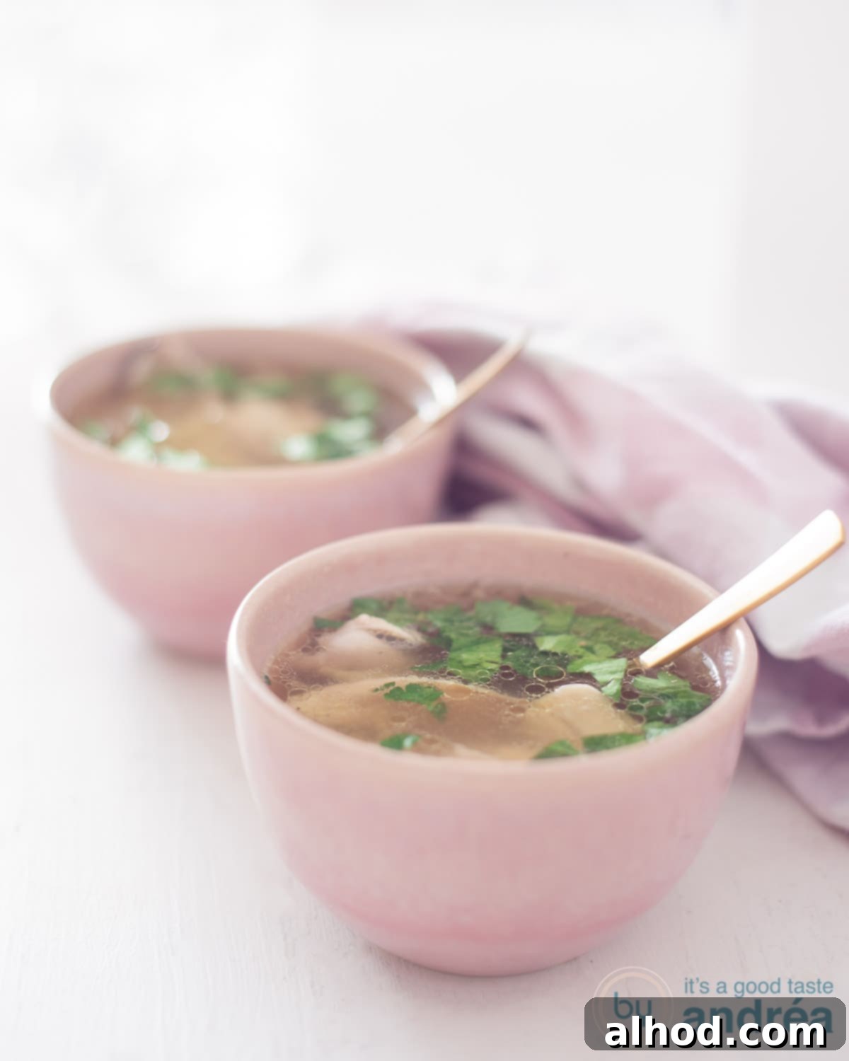 A height photo with two bowls, pink, filled with soup, lots of chicken and parsley. A white background. A pink and white tea towel.