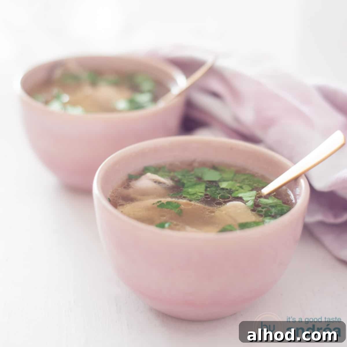A square photo with two pink bowls filled with homemade fresh chicken soup. A white background and on the right a pink and white tea towel