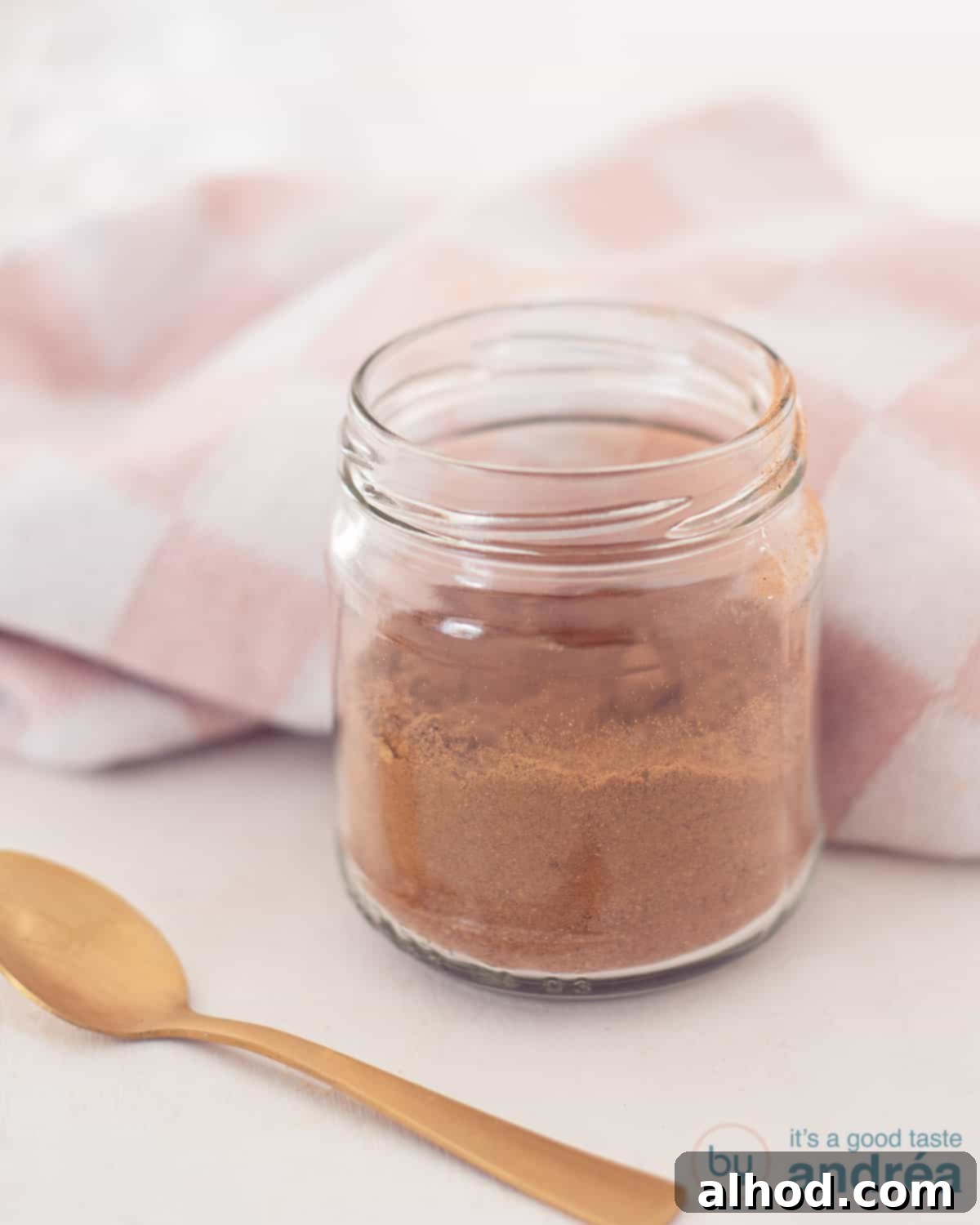 Homemade Dutch Speculaas Spice Blend 2 A glass jar filled with cookie spice mix on a white surface. A golden spoon lies in front of it. A pink and white tea towel lies behind it.