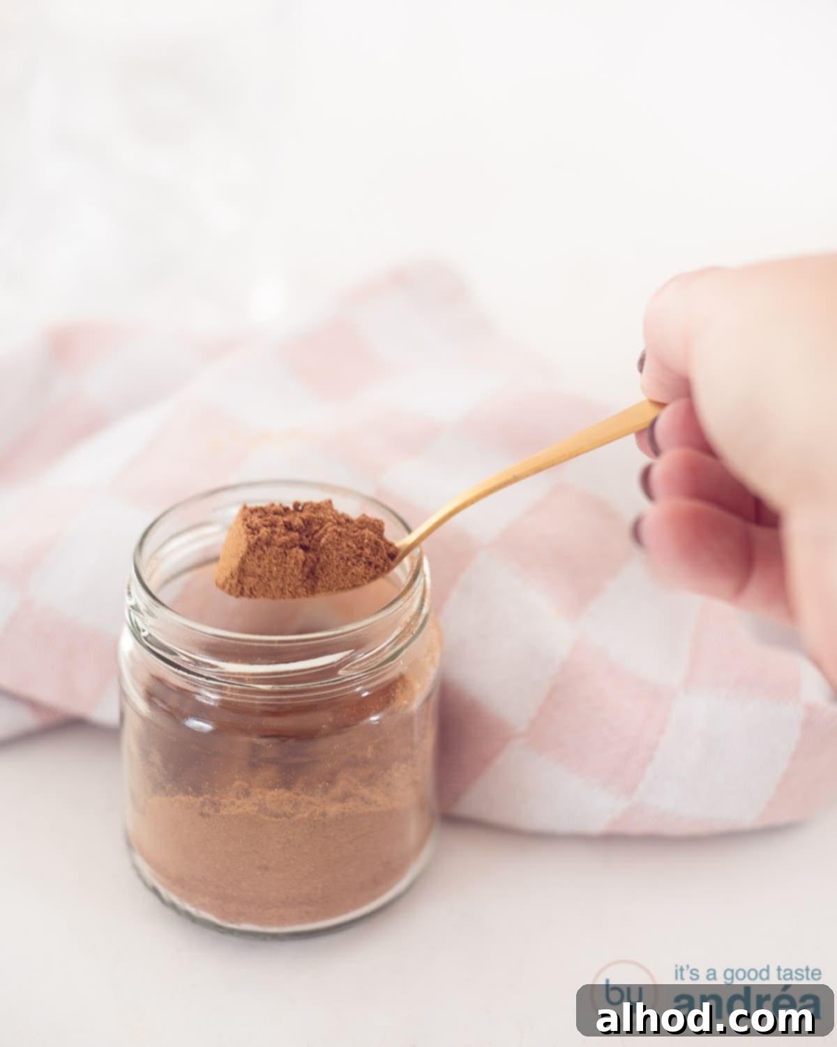Homemade Dutch Speculaas Spice Blend 3 A glass jar with gingerbread spices with a golden spoon that has some mix on the spoon. A white background. In the background a pink and white tea towel