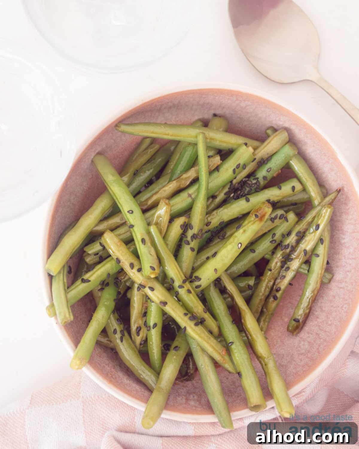 Sesame Garlic Green Beans 2 A top-down photo of a pink bowl filled with glossy, braised green beans in a rich Oriental sauce, set against a pristine white background. The beans are perfectly coated with a delicious glaze, highlighting the dish's irresistible allure.