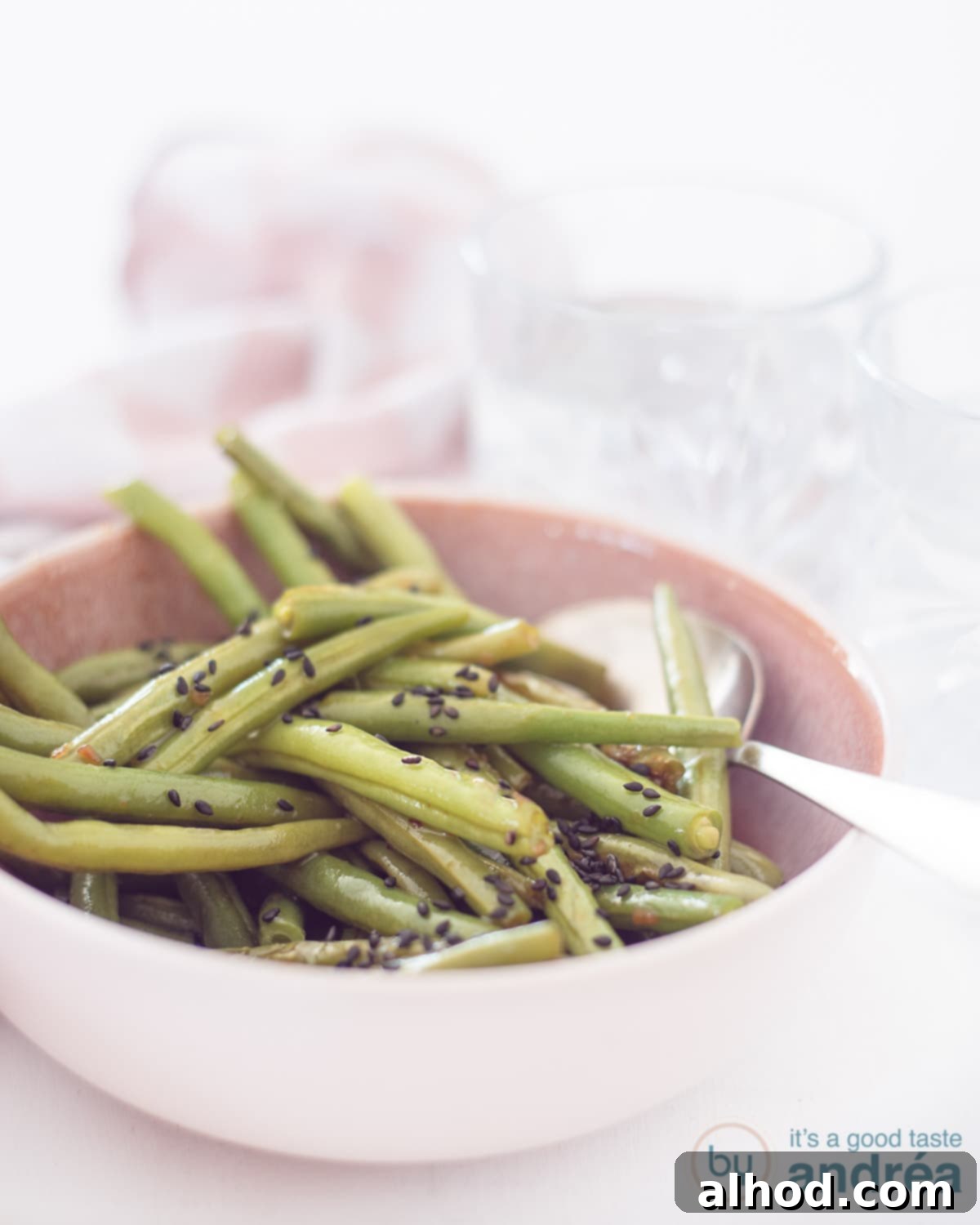 Sesame Garlic Green Beans 3 A close-up shot of a pink bowl filled with perfectly cooked, glistening Oriental green beans, complete with a serving spoon resting inside, all placed on a clean white background. The shiny sauce and fresh green color are clearly visible.