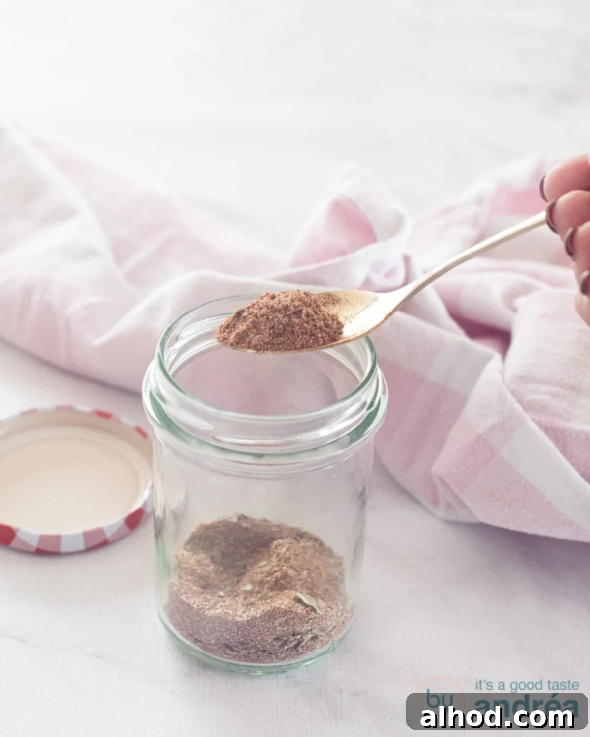 Your Go-To Homemade Chicken Seasoning 2 A glass jar with chicken spices, a golden spoon scoops out a spoonful of homemade mix. A pink and white tea towel in the back.