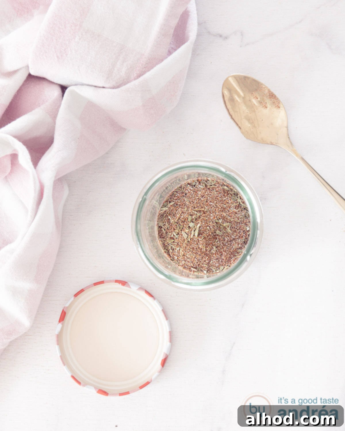 Your Go-To Homemade Chicken Seasoning 3 A photo from above showing a glass jar with homemade chicken seasoning. A white marble surface. A pink and white tea towel at the top left