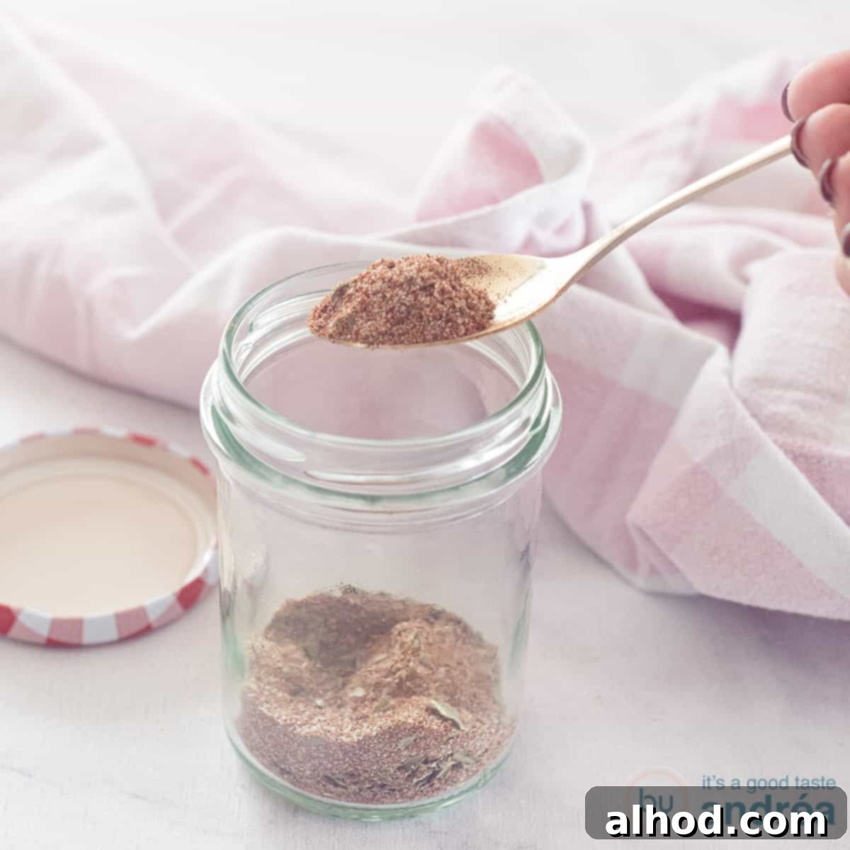 Your Go-To Homemade Chicken Seasoning 4 A square photo with a glass jar with chicken spices that are mixed themselves. A spoonful of mix is scooped out. A pink white tea towel in the background.