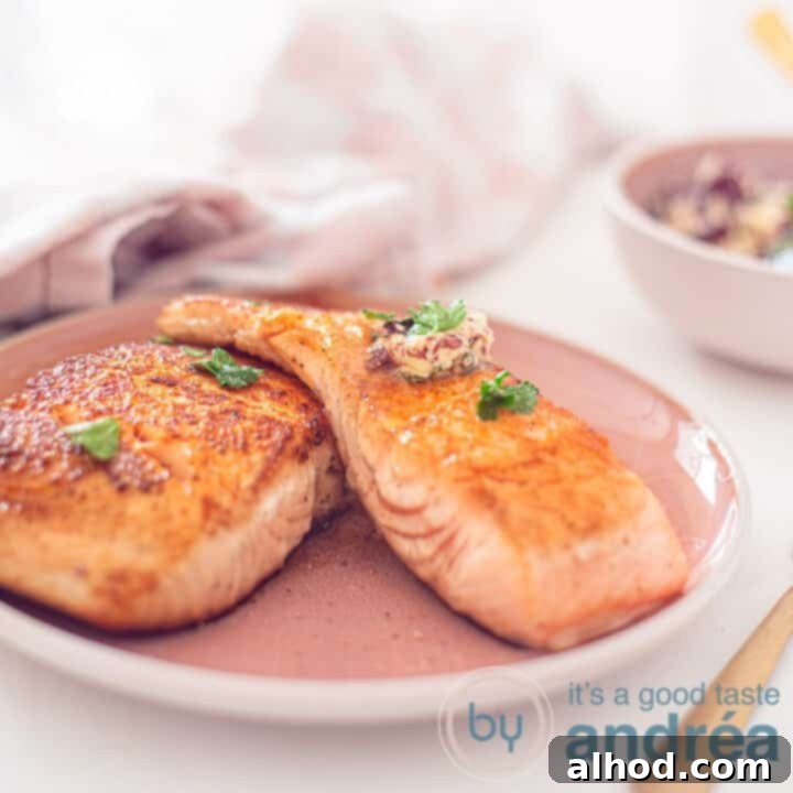 A square photo of a pink plate on a white background. Two baked salmon fillets with red wine herb butter on the plate. In the background a pink and white tea towel.