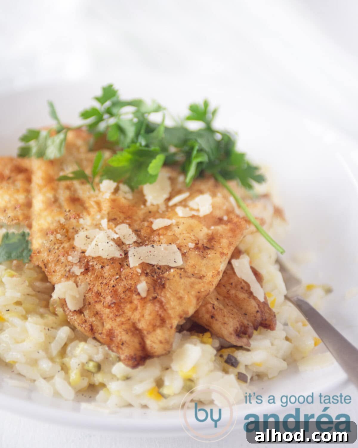 Photo of a white plate with a gold spoon. The plate is filled with risotto with zucchini and cheese. On top of that are two slices of fried cod