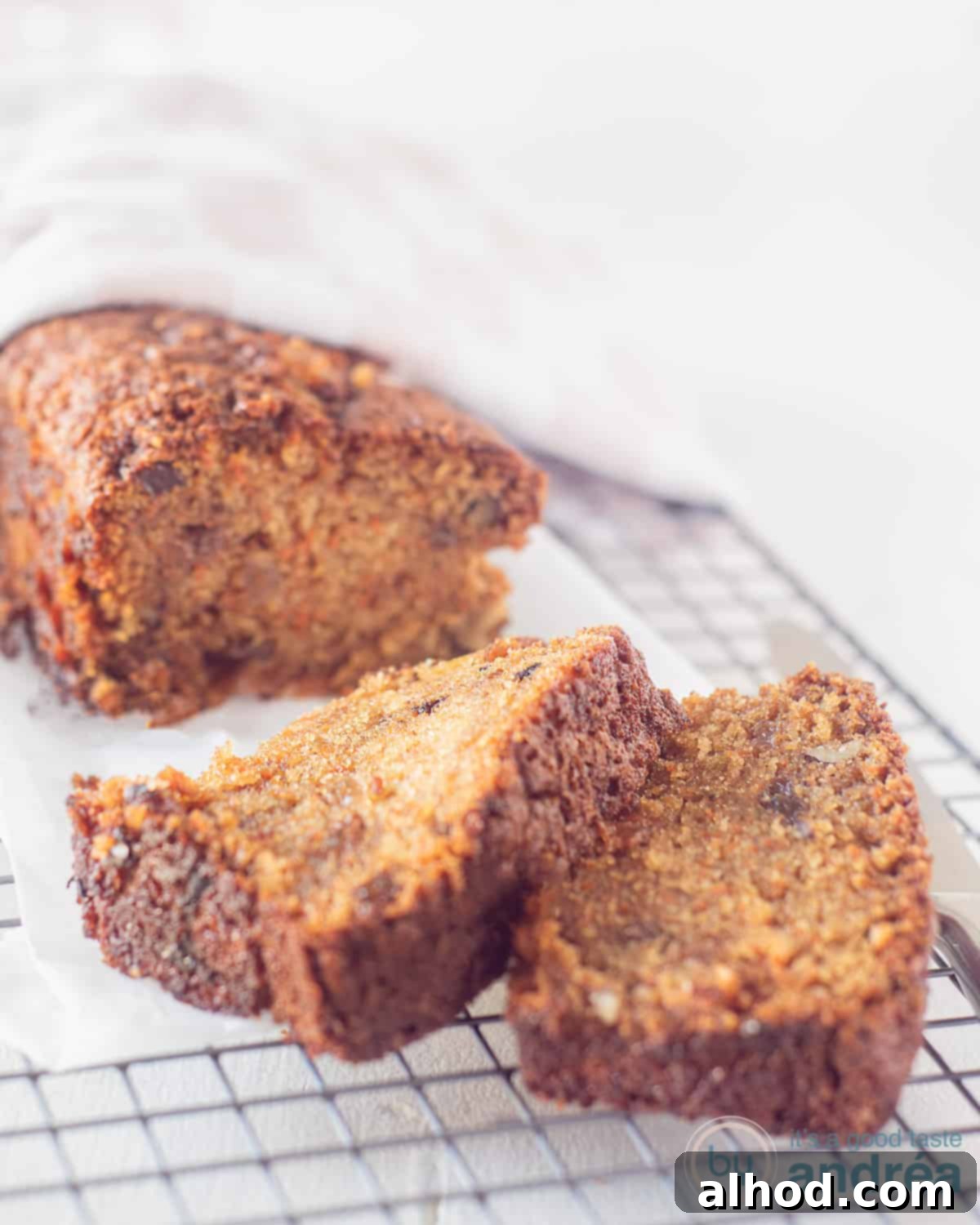 Spiced Carrot Raisin Pound Cake 2 A beautifully baked carrot cake, rich with raisins and walnuts, resting on a cooling rack over a baking sheet, partially covered by a charming pink and white tea towel. Two enticing slices are cut and placed in front, ready to be enjoyed.
