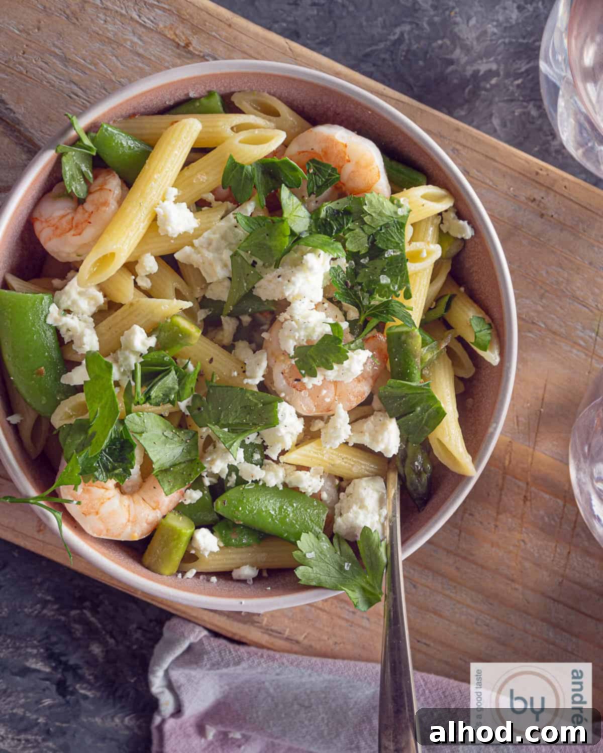 Shrimp Primavera with Spring Greens 2 A photo from above of a pink bowl with pasta, shrimp, spring vegetables, feta and parsley. Everything on a wooden board.