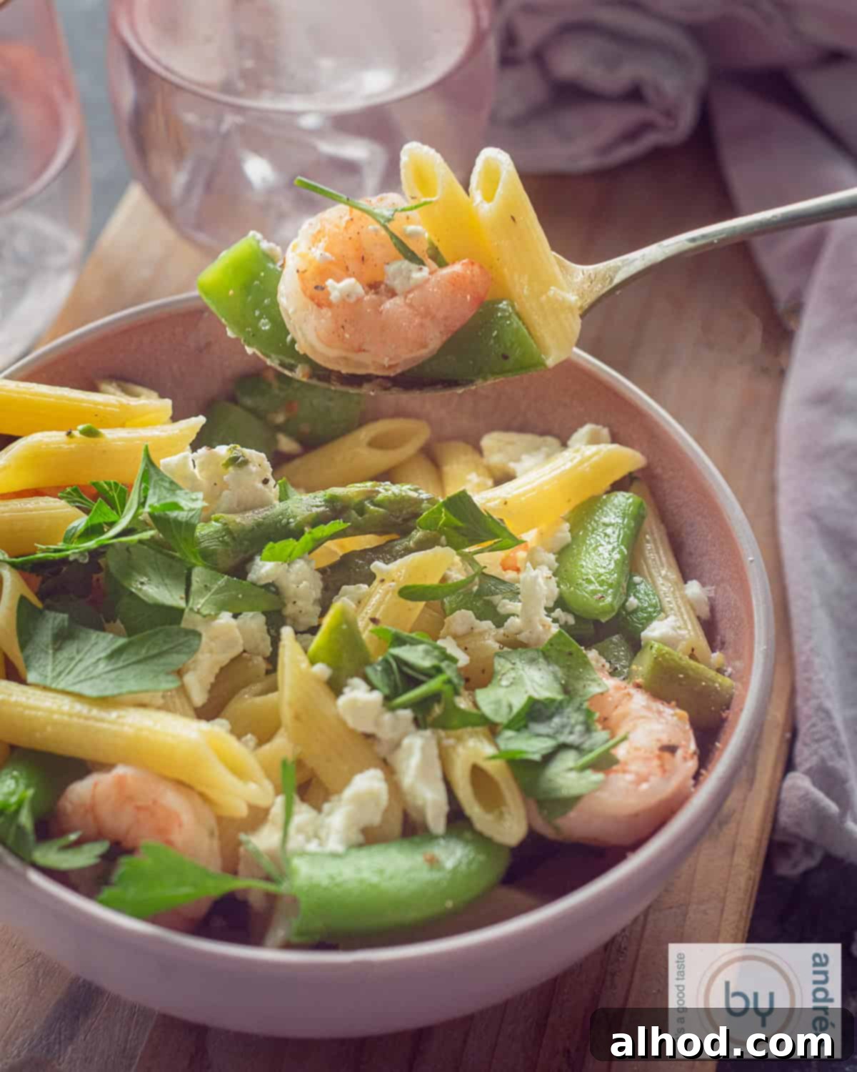 Shrimp Primavera with Spring Greens 3 An overhead photo with a pink bowl on a wooden board filled with pasta, shrimp, sugar snap peas and asparagus. A spoon scoops out a bite.