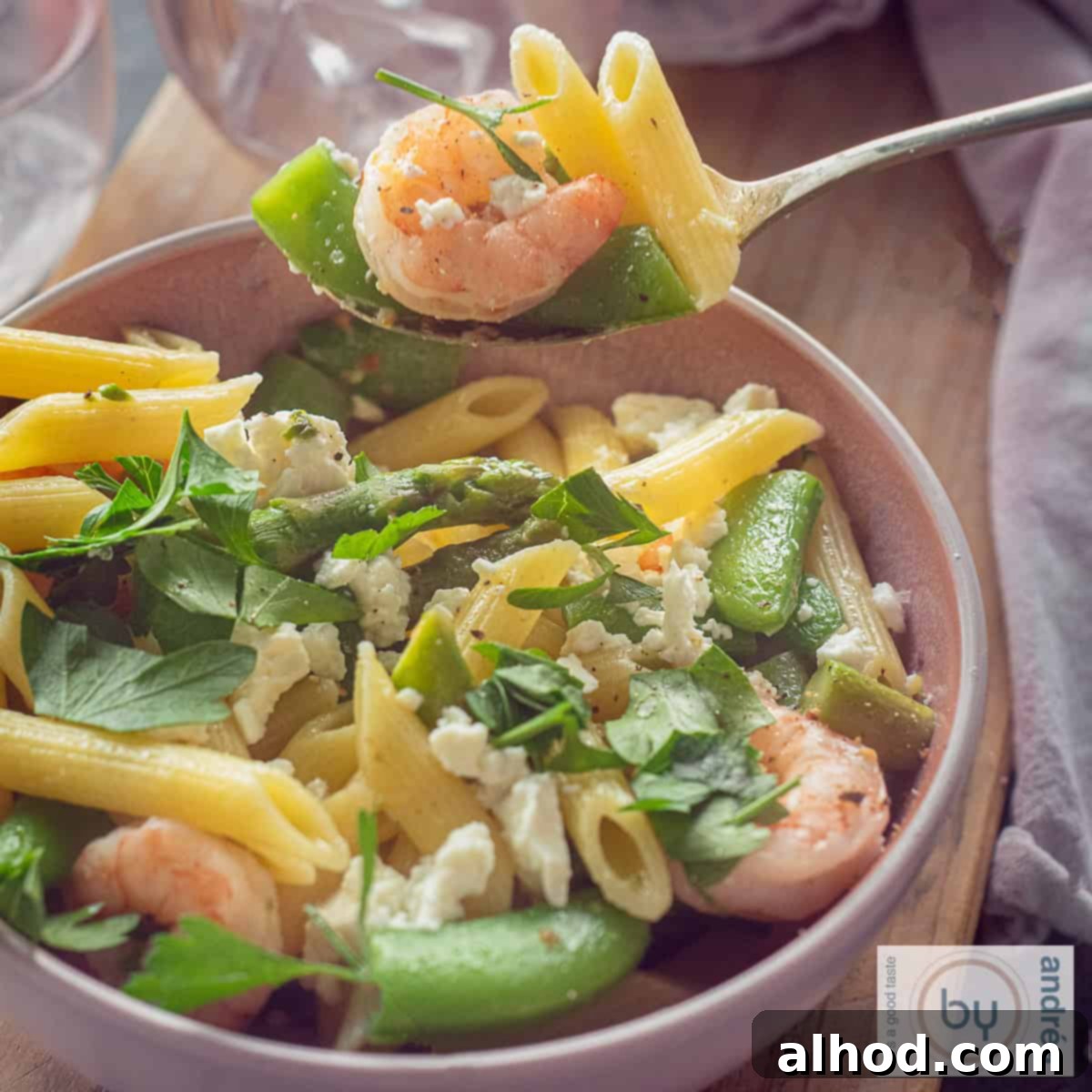 Shrimp Primavera with Spring Greens 4 A square photo of a pink bowl with a mix of shrimp, penne and spring vegetables. A spoon scoops out a bite.