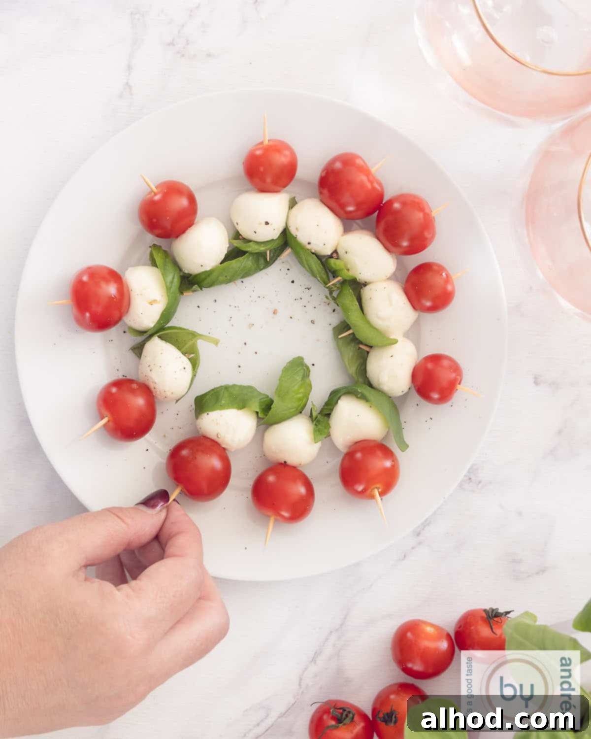 Overhead view of a white platter brimming with mini Caprese skewers, as a hand reaches to pick one.