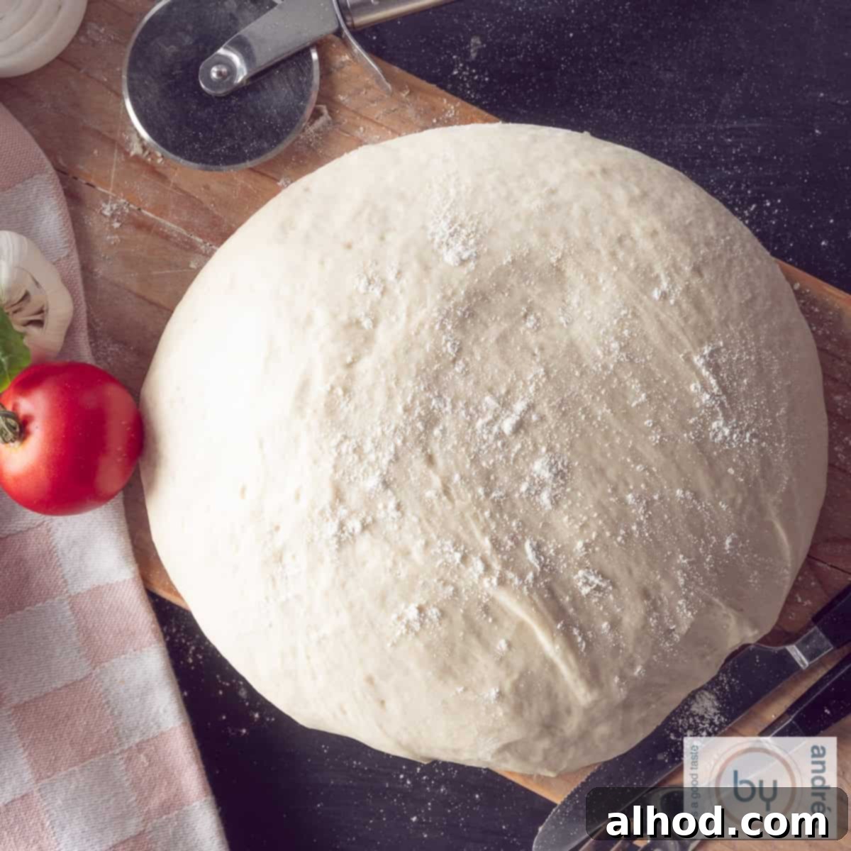Effortless Homemade Pizza Dough 6 A square photo taken from above of a ball of pizza dough on a wooden board. A tomato and garlic and a pizza cutter are next to it.