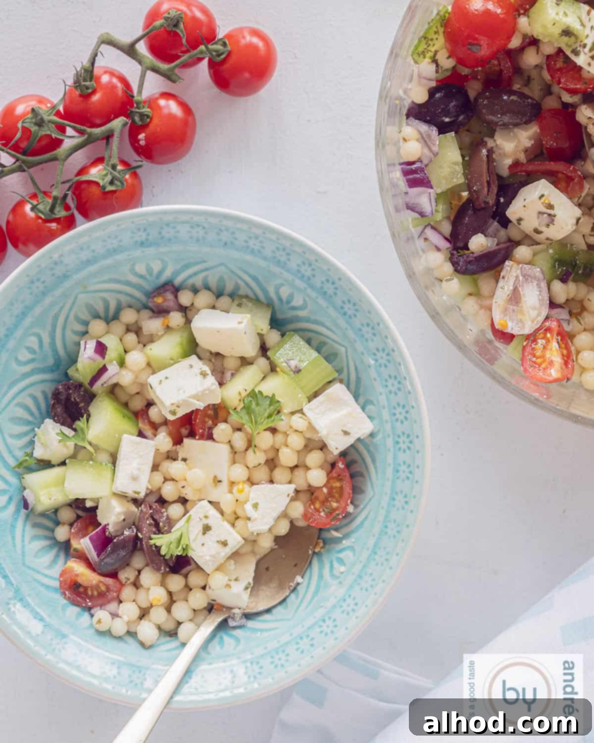 A photo from above of a blue and a glass bowl filled with pearl couscous salad with Mediterranean vegetables and feta. A bunch of tomatoes above