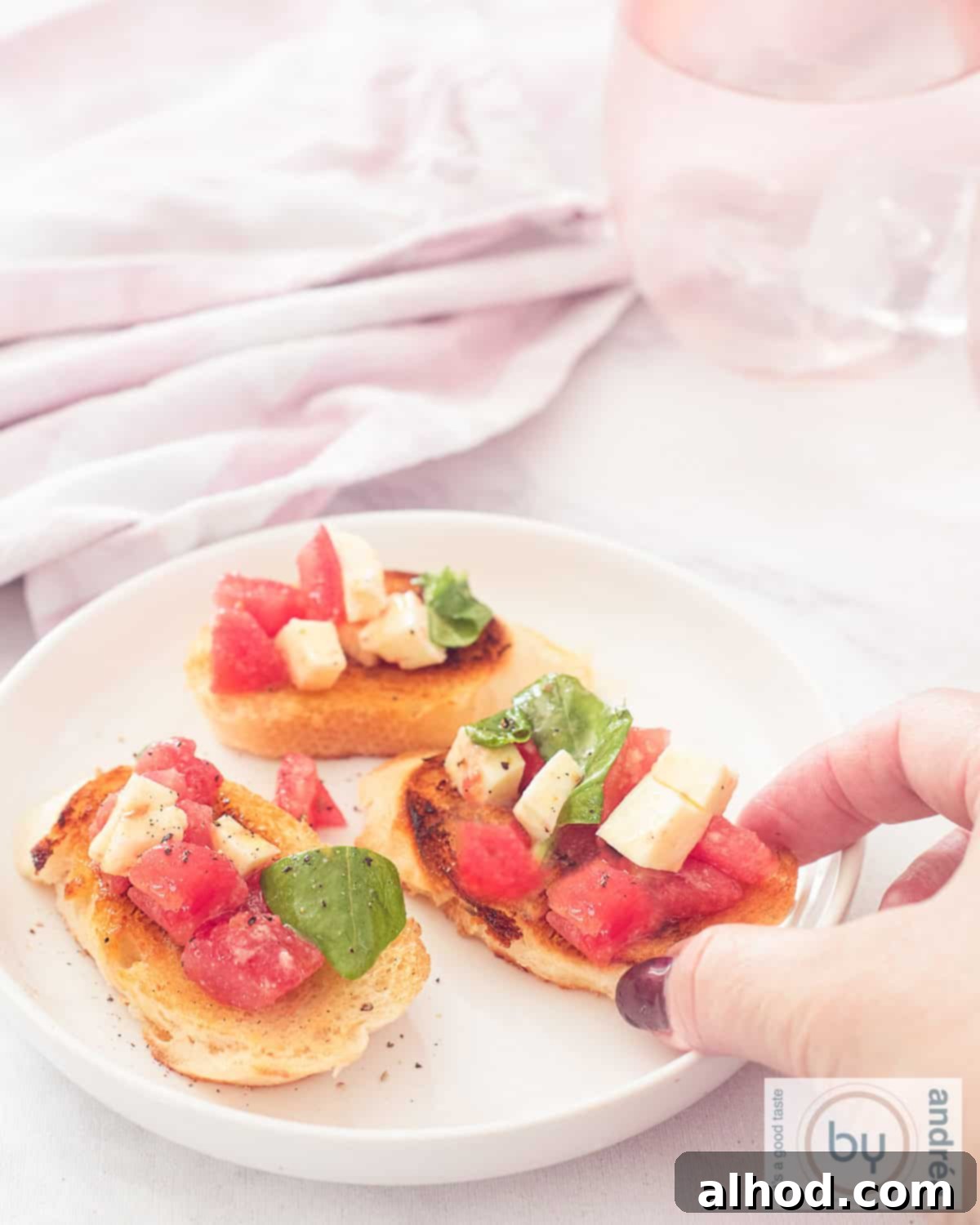 An aerial photo of a white plate with three bruschetta baguettes with tomato, mozzarella and basil topping. A hand takes a bruschetta.
