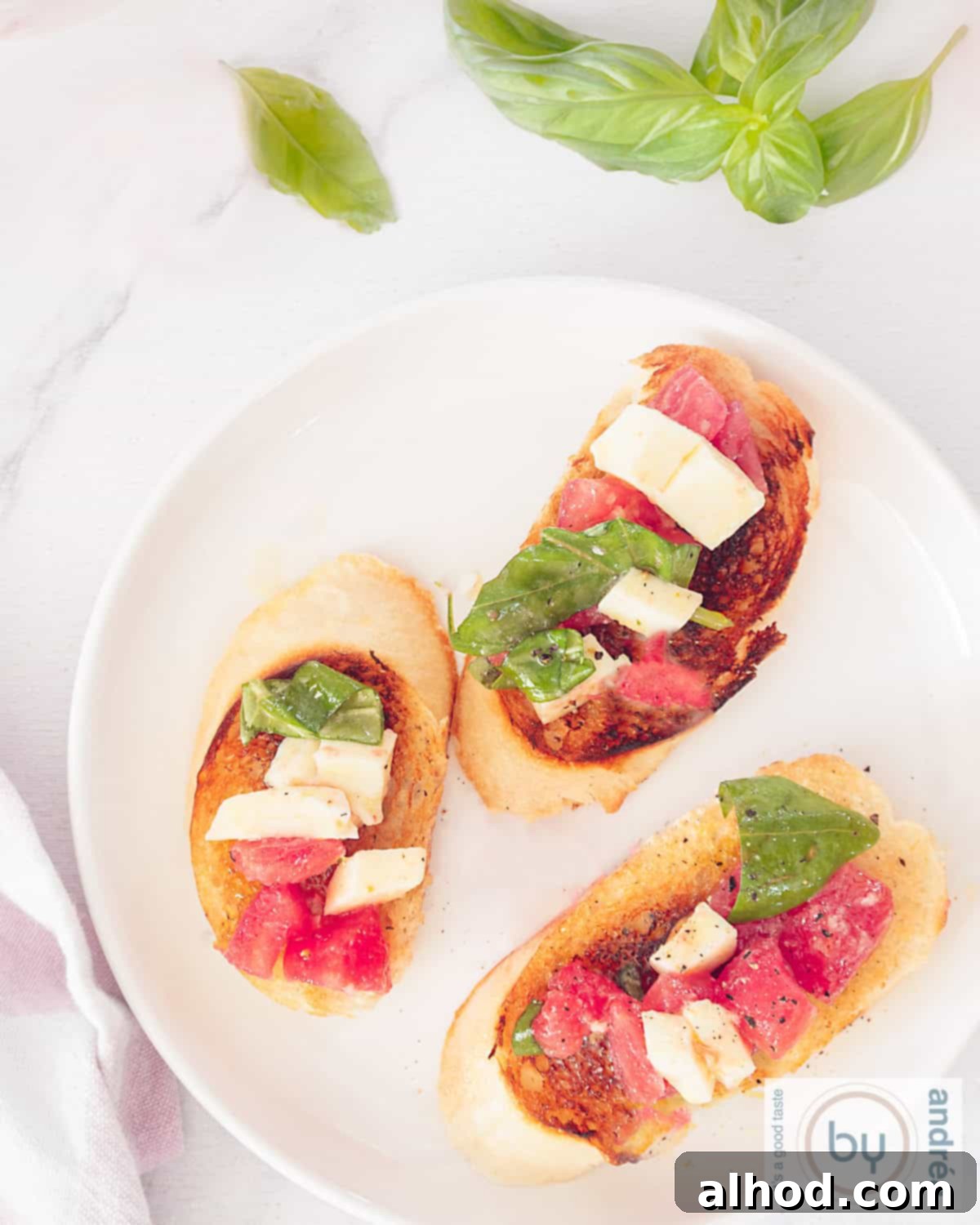A top-down photo of a white plate with three pieces of grilled baguette with a tomato mozzarella salad. In the top soil basil leaves.