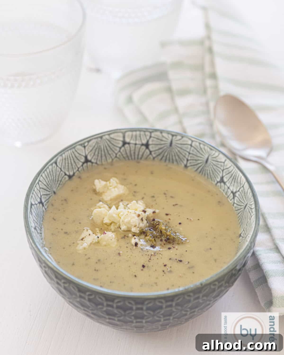 A green bowl with creamy zucchini soup, topped with fresh feta crumbles and a swirl of green pesto, set against a pristine white background. To the right, a charming green and white striped tea towel lies next to a golden spoon.