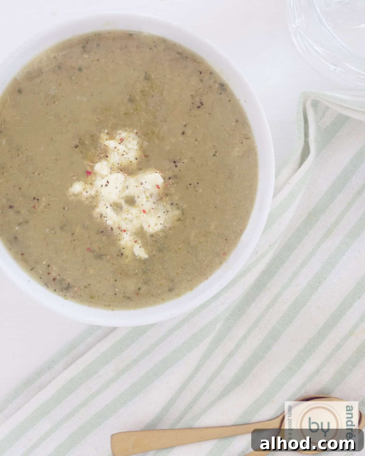 Velvety Zucchini Soup with Bright Pesto and Feta 5 A high-angle photograph of a white bowl filled with creamy zucchini soup, adorned with feta cheese and a swirl of pesto. A green and white striped tea towel is neatly folded in the bottom left corner.