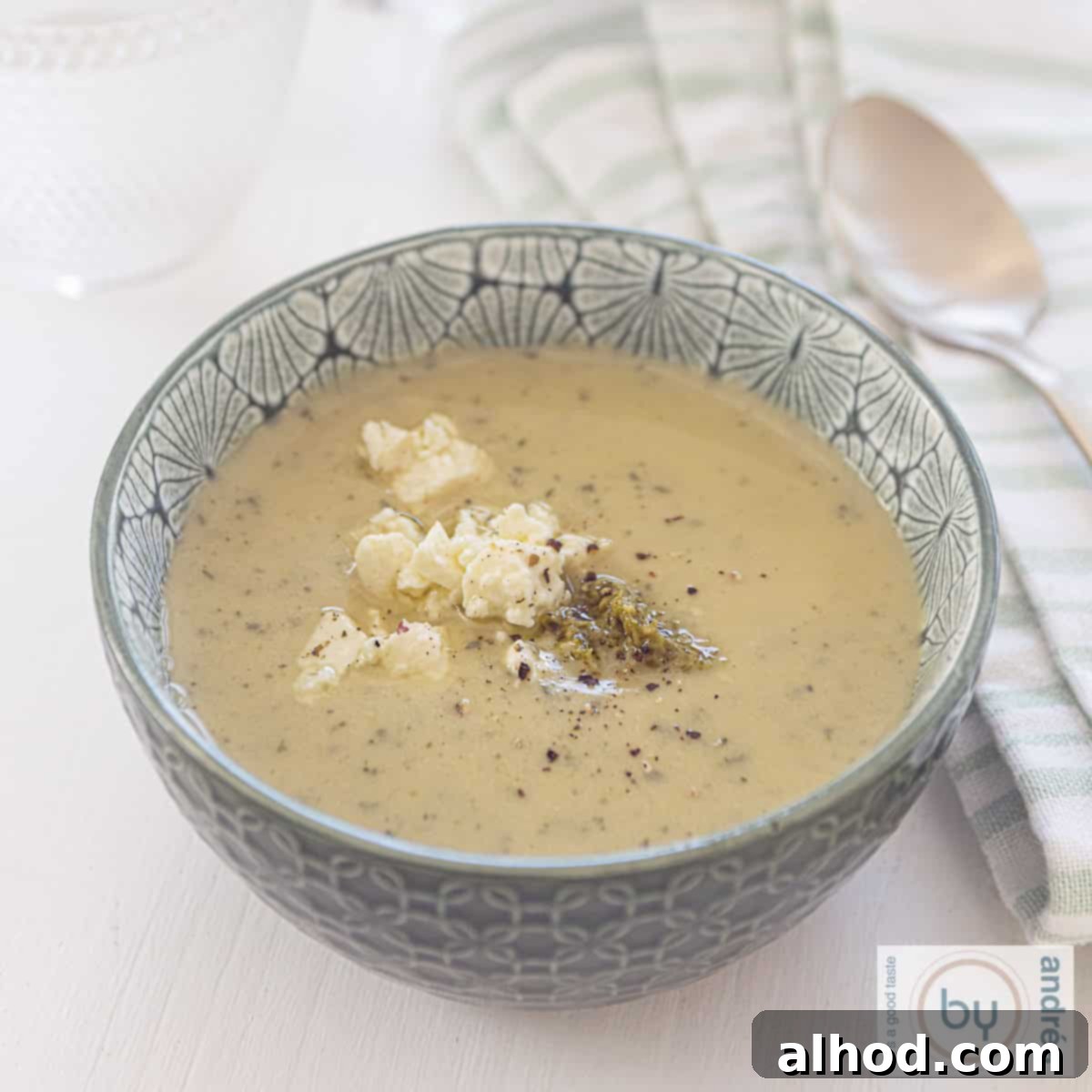Velvety Zucchini Soup with Bright Pesto and Feta 6 A square photo with a white background, a green bowl in the middle with courgette soup with cream, feta and pesto. A green and white tea towel is on the right.