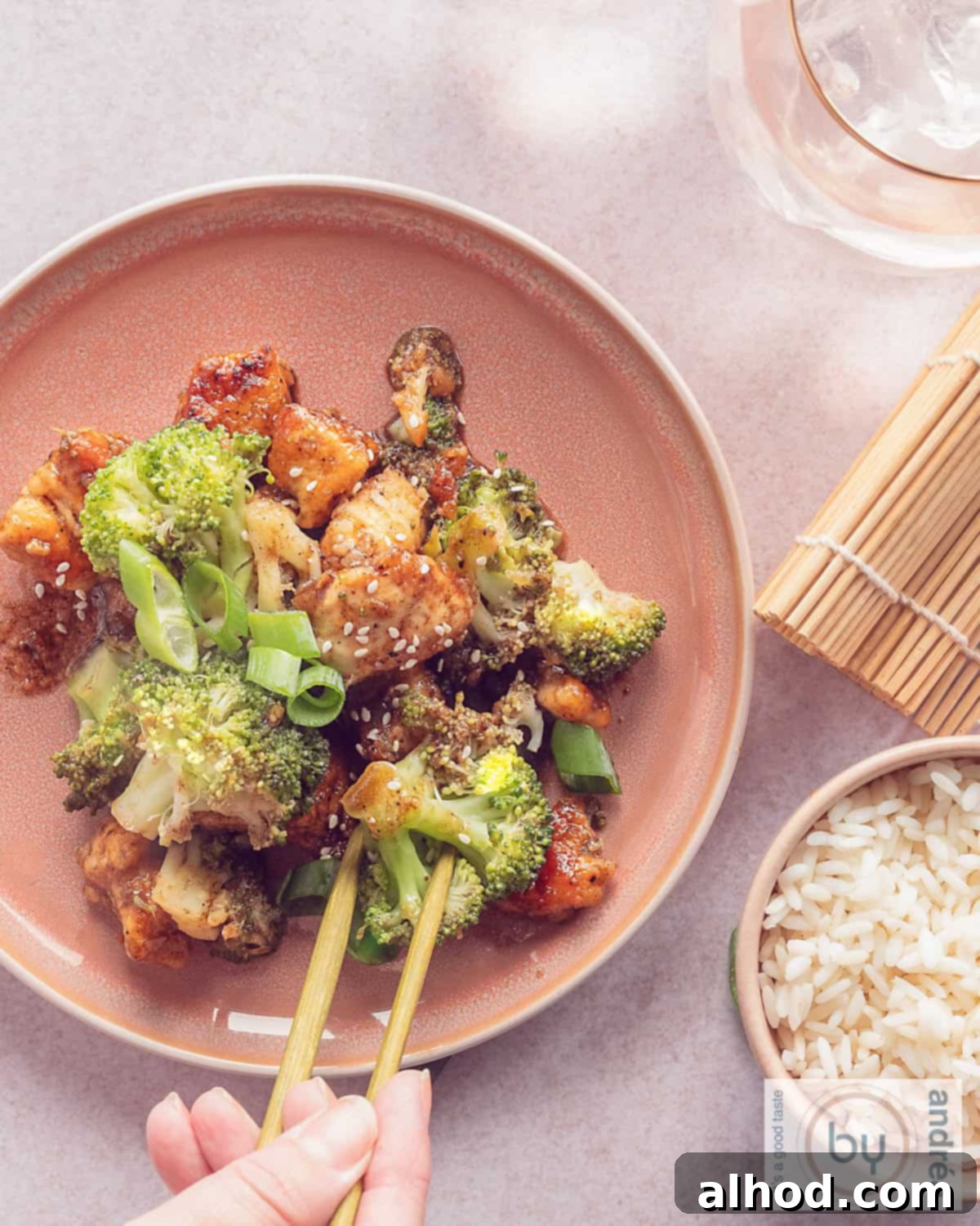Easy Hoisin Chicken and Broccoli Stir Fry 5 A photo from above a pink plate with broccoli, chicken stir-fried in Asian sauce. Two chopsticks grab a piece. A bowl of rice bottom left.