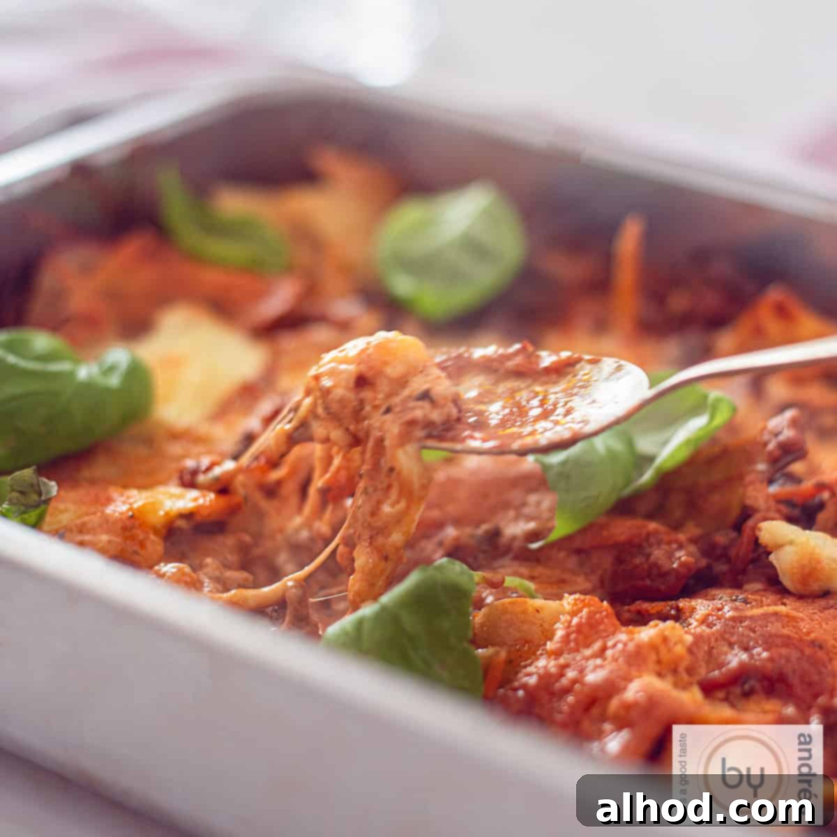Decadent One Pot Bacon Mascarpone Lasagna 6 A square photo of a roasting pan filled with cheesy one-pan lasagna with mascarpone and minced meat. A spoon takes a bite.