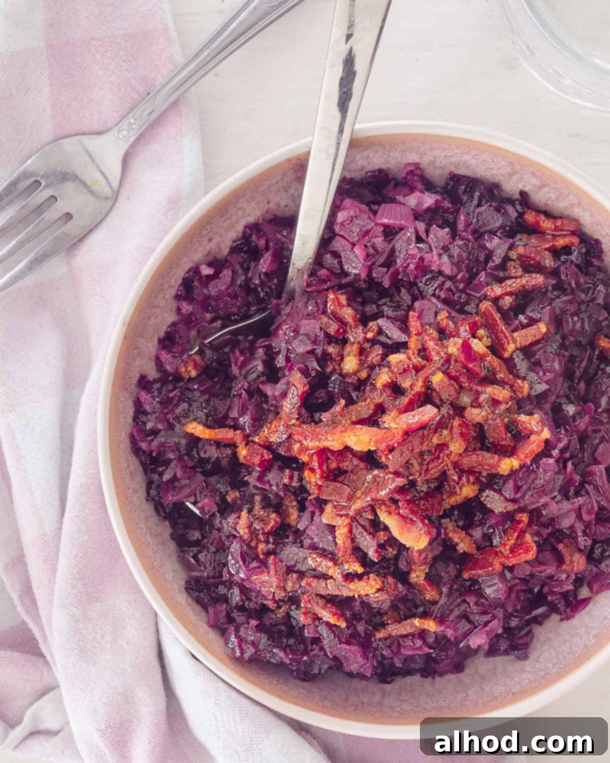 Sweet Sour and Savory German Red Cabbage with Bacon and Apples 5 A photo from above of a pink bowl with red cabbage and a topping of bacon. A white background and on the left a pink and white tea towel.