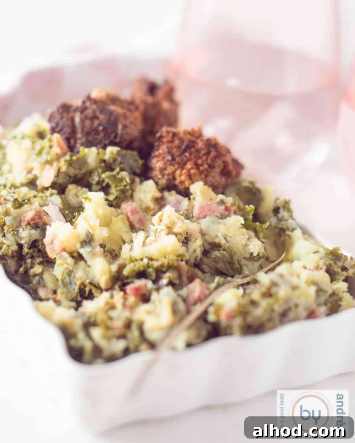 An height photo of a white bowl with boerenkoolstamppot (kale mash) with bacon and meatballs in gravy. Pink glasses in the background