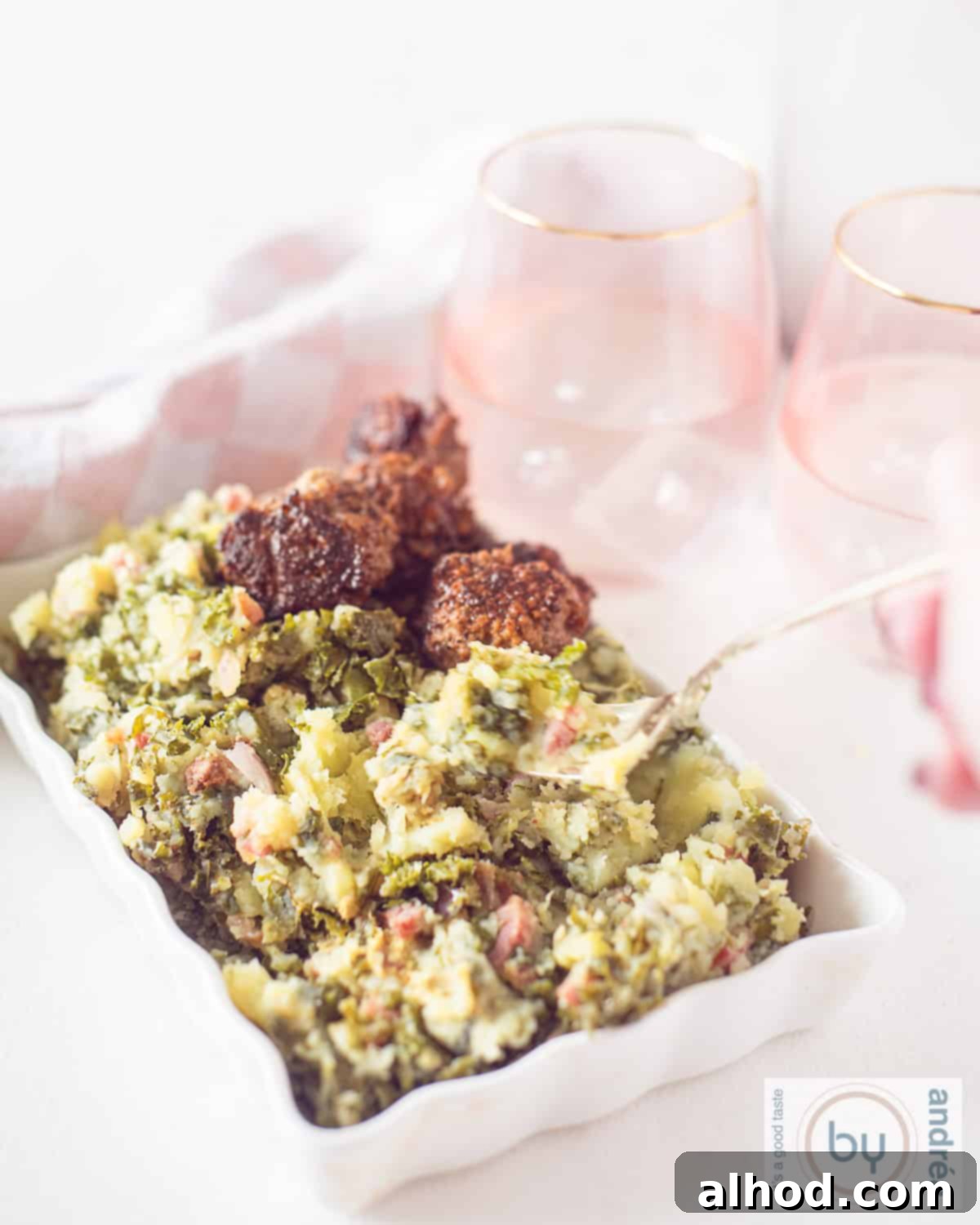 An aerial shot of a white oven dish with kale mash and meatballs. A fork pokes a bite out of it. Pink glasses in the background.