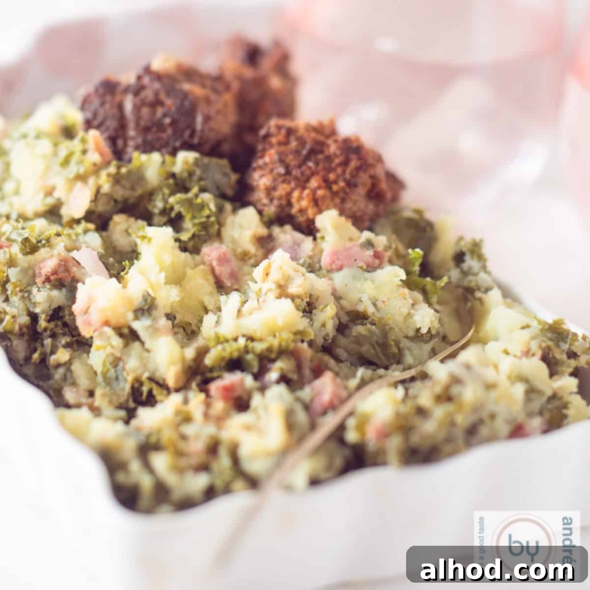 A square photo of a white bowl filled with kale mash, bacon, meatballs and gravy.