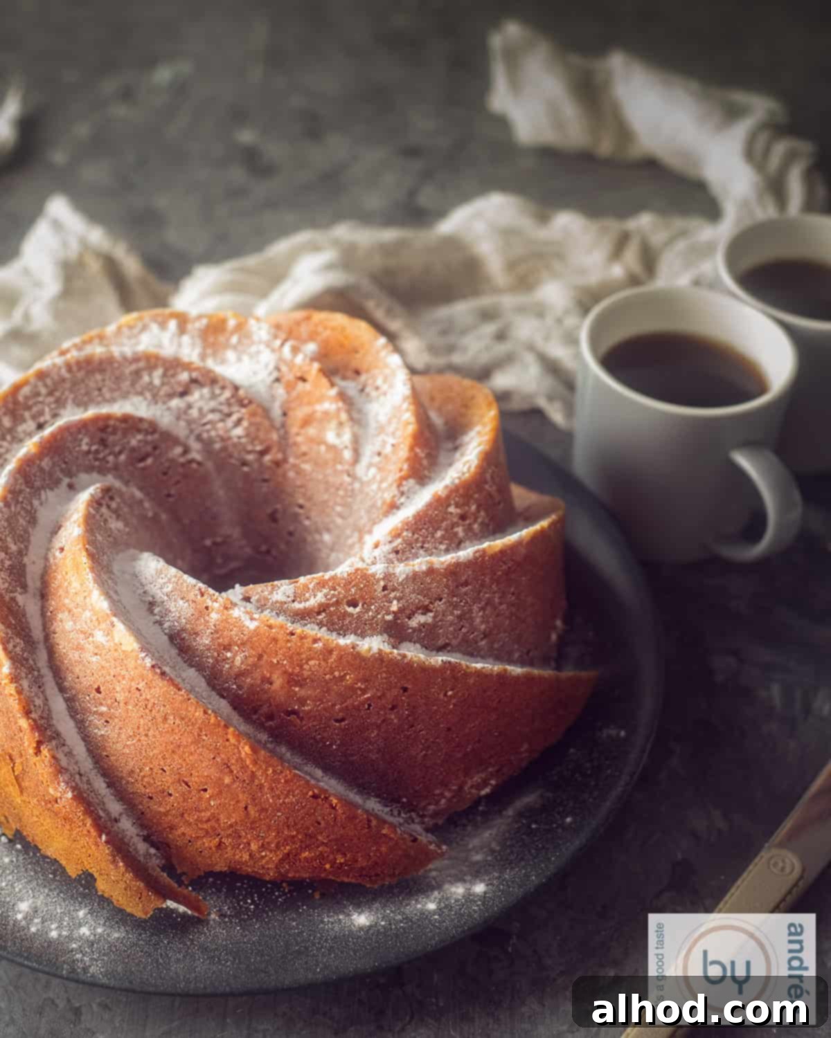An elevation photo of a pumpkin bundt cake on a gray plate. Two cups of coffee behind it, a dark background.