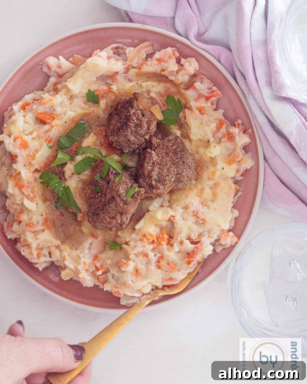 Velvety Dutch Hutspot with Zesty Meatballs 5 A top-down photo featuring a white plate with creamy Hutspot and three spicy meatballs, generously covered in rich gravy, all on a clean white background. A pink and white tea towel is visible in the top right corner.