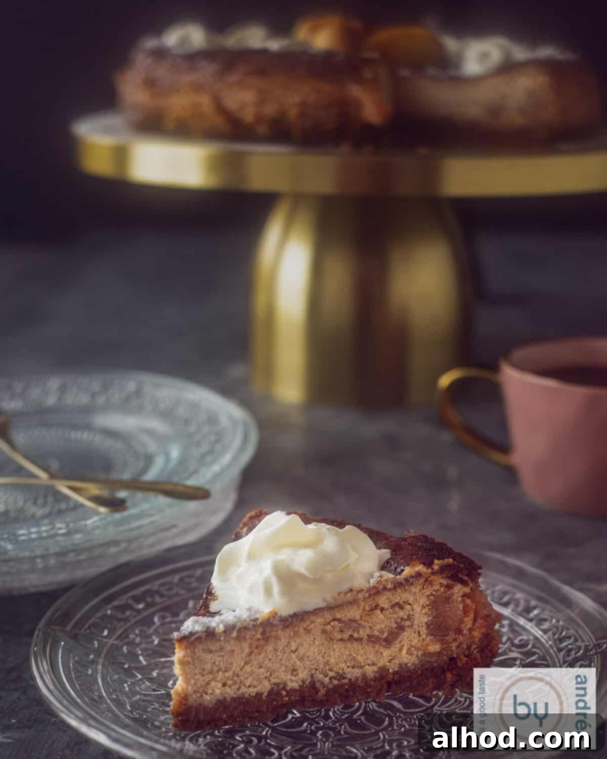 Decadent Caramel Apple Biscoff Cheesecake 5 A piece of caramelized apple cheesecake on a glass plate with a dollop of whipped cream. In the background a golden cake stand with the rest of the cheesecake.