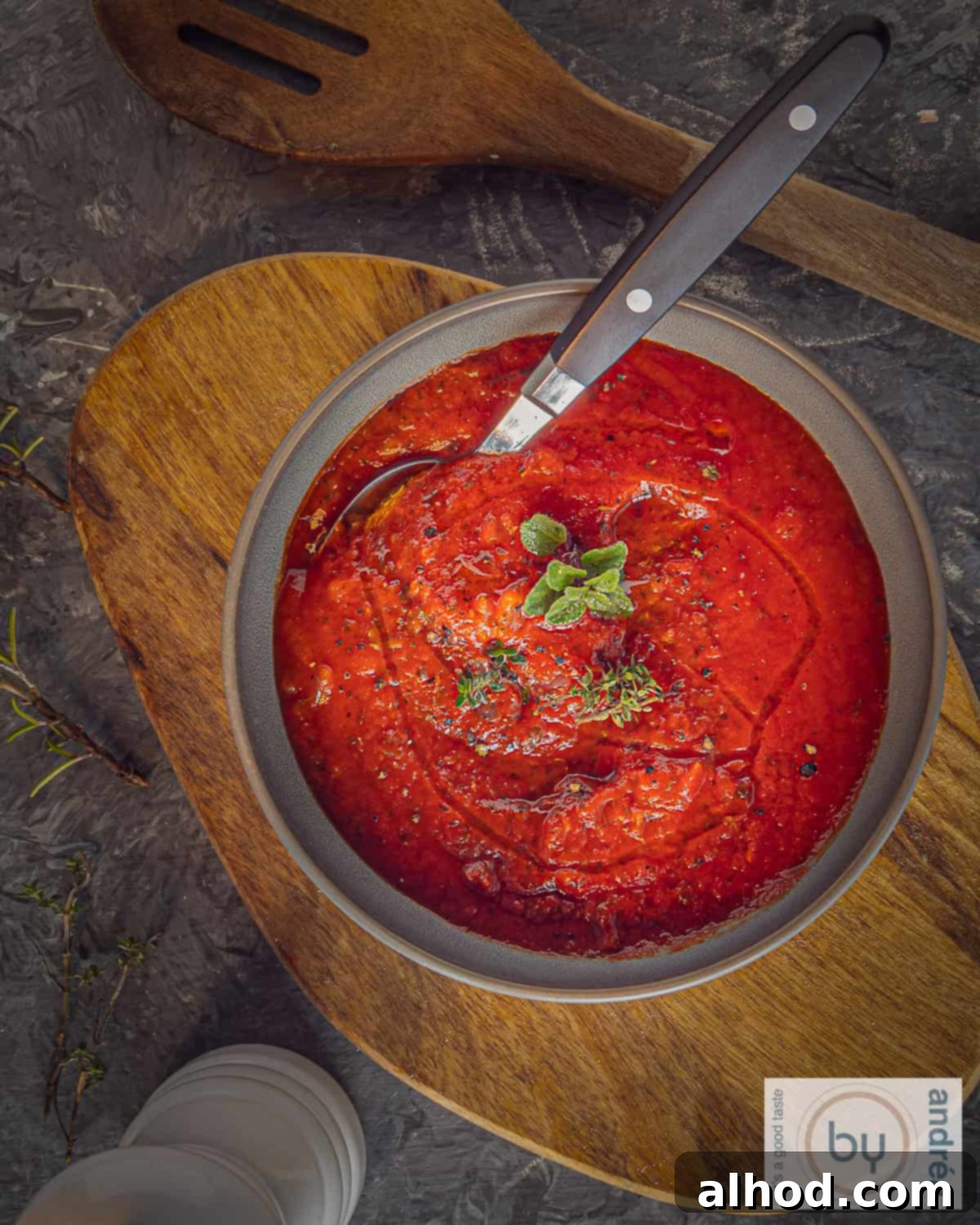A top-down photo of a wooden board with a gray bowl filled with pizza sauce on it. A spoon is in the bowl.