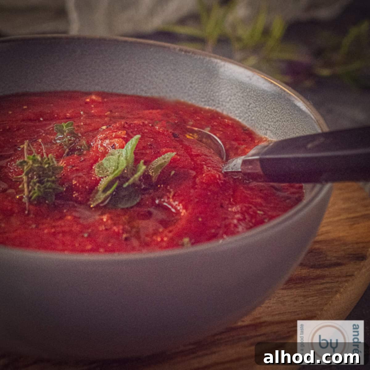 A square photo showing part of a gray bowl filled with pizza sauce with fresh herbs. A spoon is sticking in it.