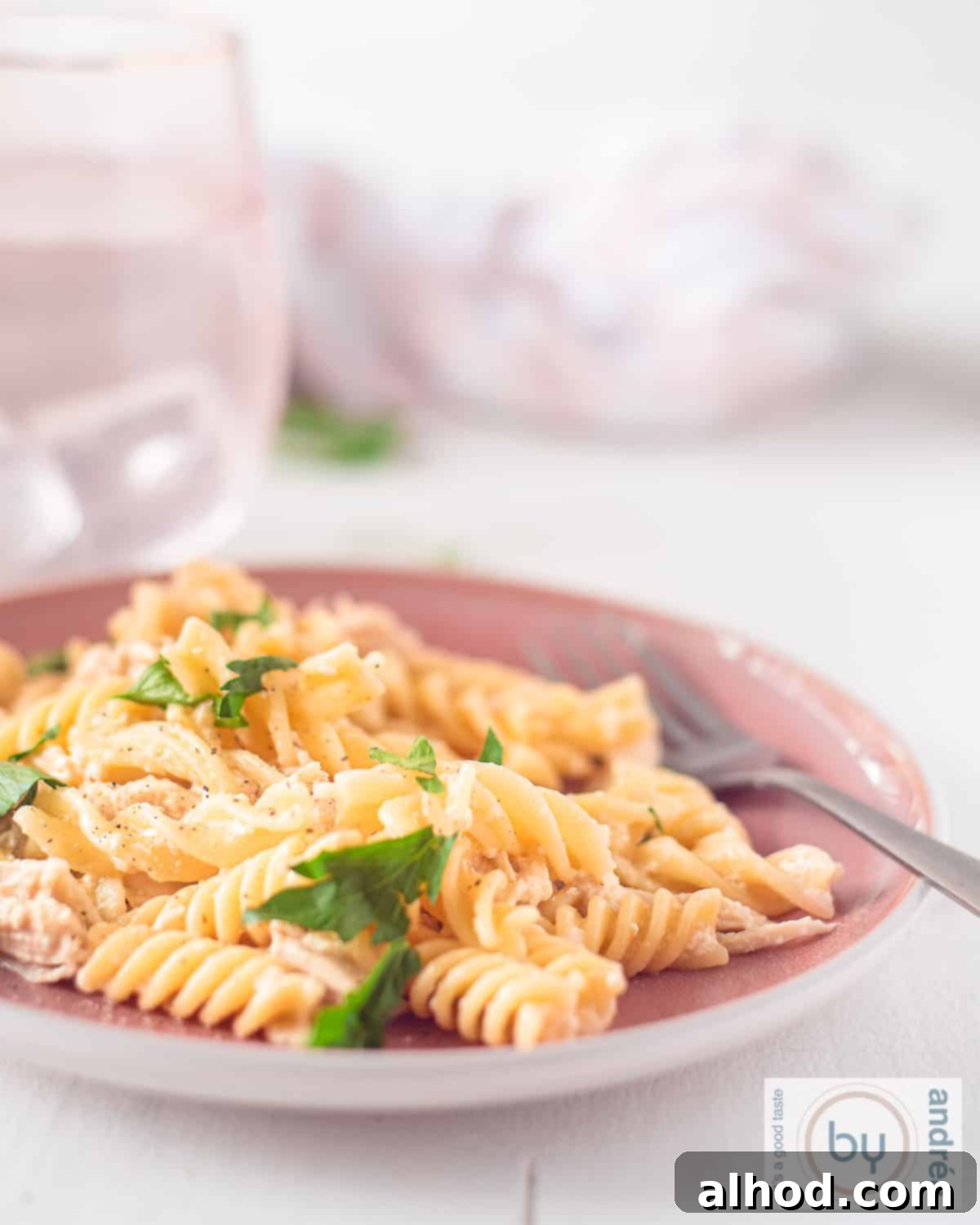 Creamy Garlic Parmesan Chicken and Fusilli in the Slow Cooker 5 An aerial photo of a pink plate filled with fusilli pasta, creamy chicken, garlic parmesan sauce, and a garnish of fresh parsley. In the background, pink glasses with water are visible.