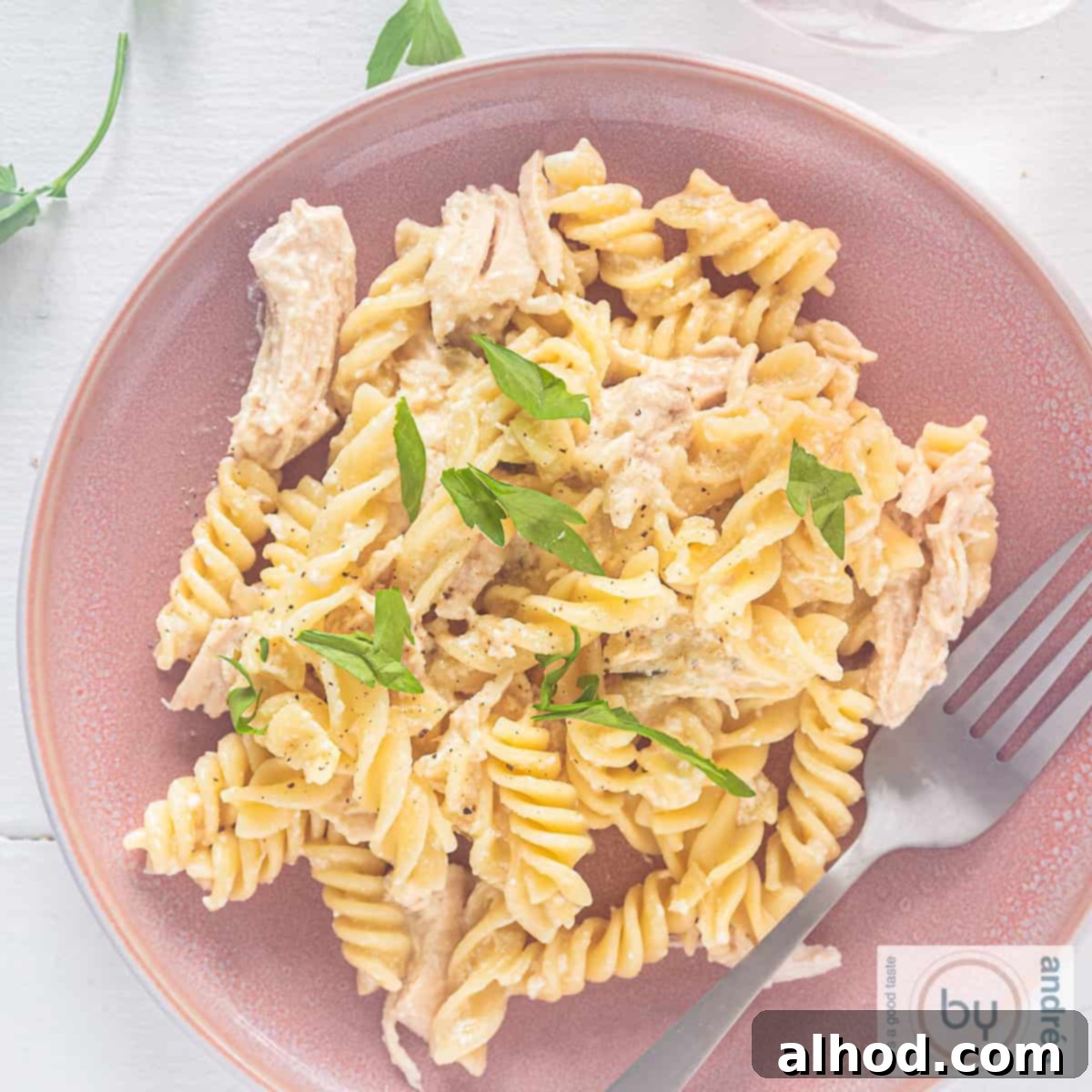 A square photo of a pink plate with fusilli pasta, creamy garlic Parmesan chicken sauce, and fresh parsley on a white background.