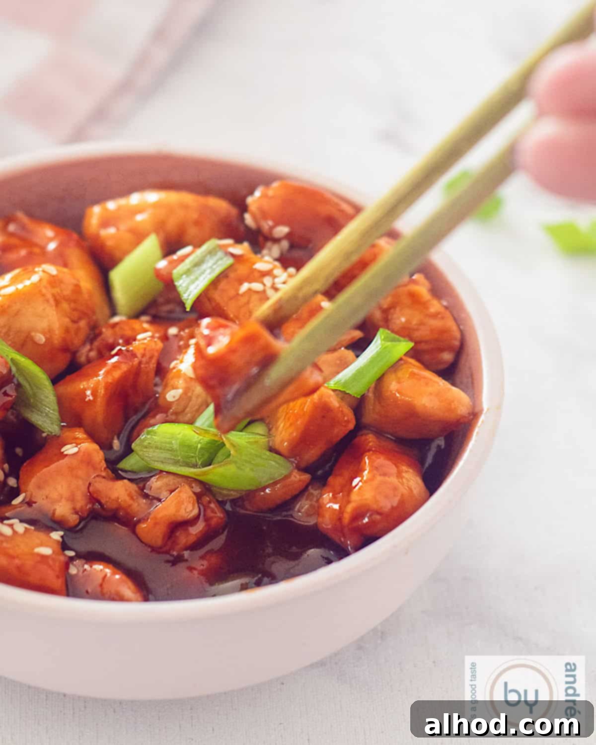 Irresistible Sweet and Sticky Garlic Chicken 5 a portion of a pink bowl of sticky chicken with a garnish of spring onions and sesame. Two chopsticks grab a piece of chicken.