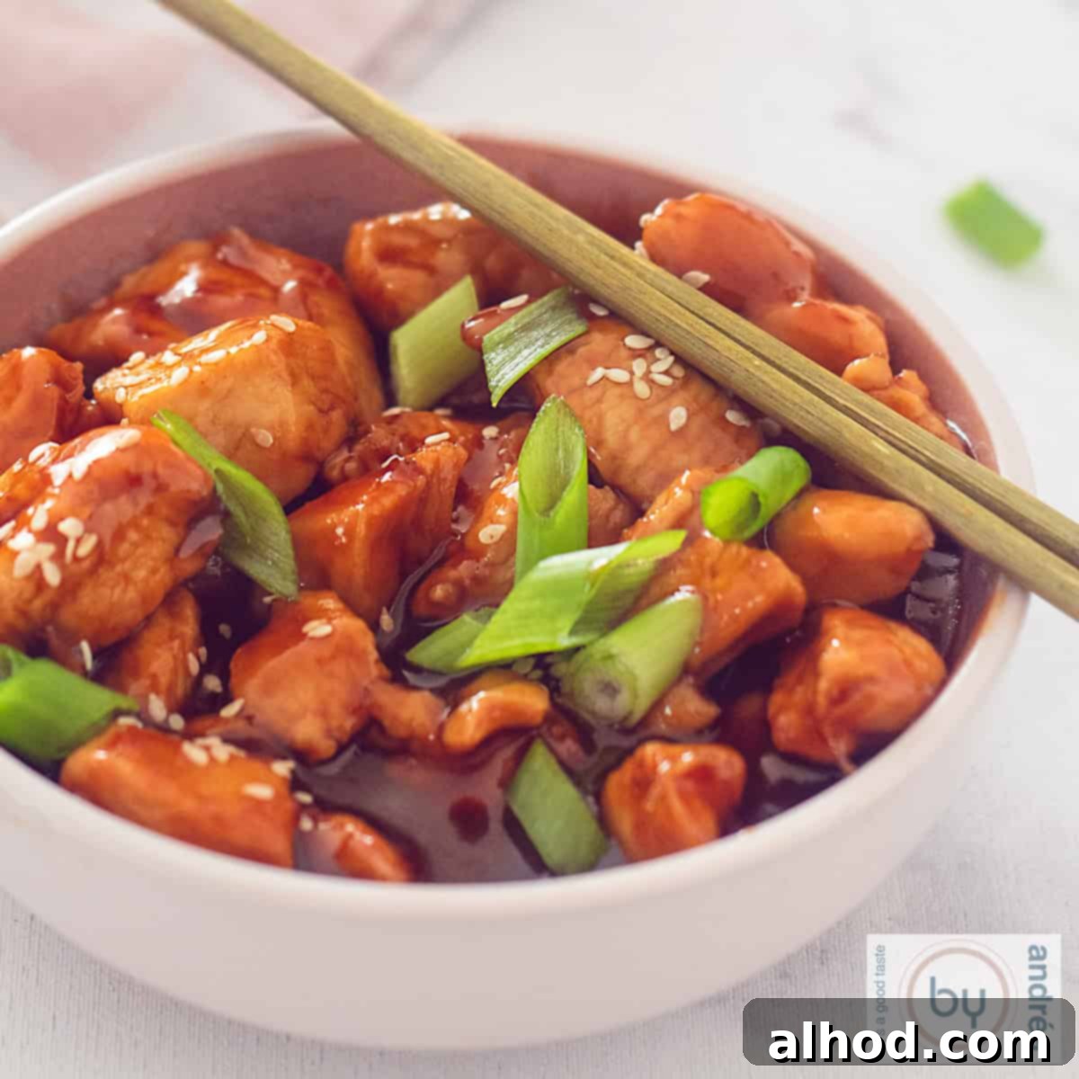 Irresistible Sweet and Sticky Garlic Chicken 6 A square photo of a pink bowl filled with chicken, sticky sauce, spring onion and sesame. Two chopsticks are on top.
