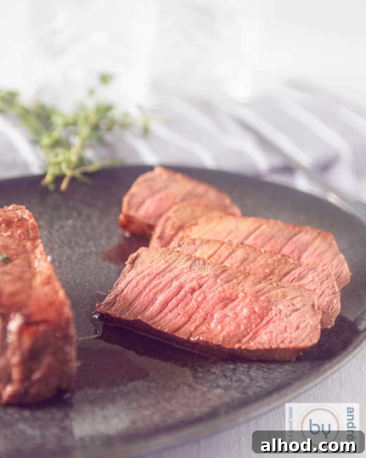 An elevation photo of roast beef, where five slices have been cut off on a grey platter. Some fresh herbs on top.