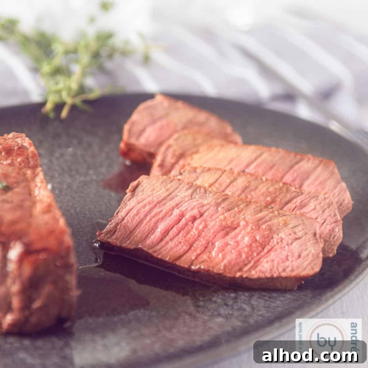 Succulent Roast Beef with Aromatic Herb Garlic Butter 6 A square photo of roast beef, with slices cut off on a grey platter. Fresh herbs and a grey white cloth in the background.