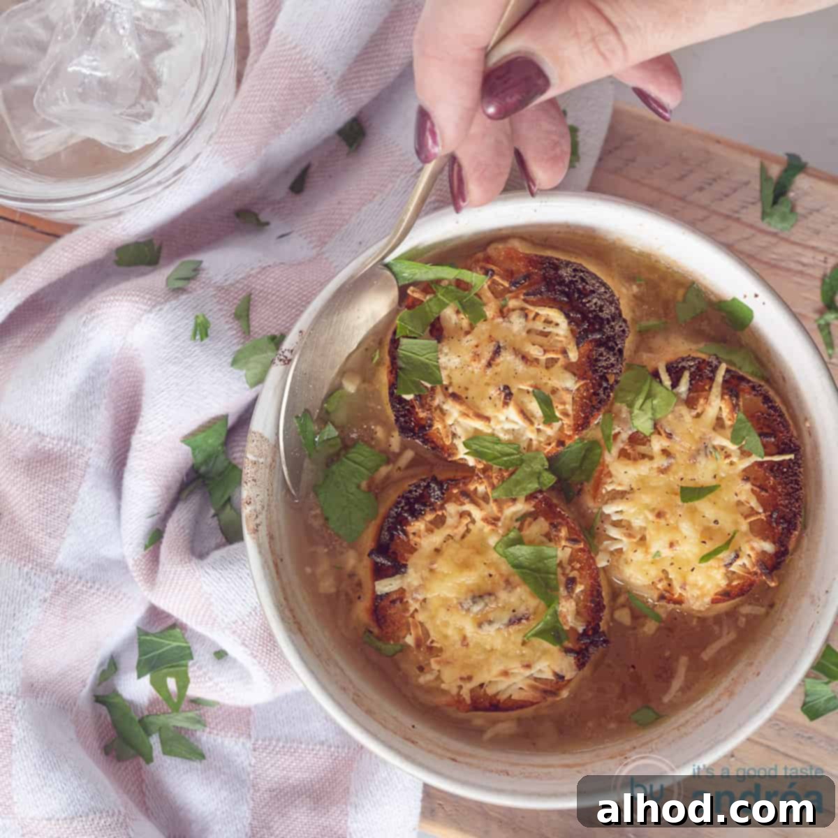 A photo from above of a white bowl of onion soup with three pieces of gratinated baguette. A spoon takes a bite. A pink and white tea towel is next to it on the left.