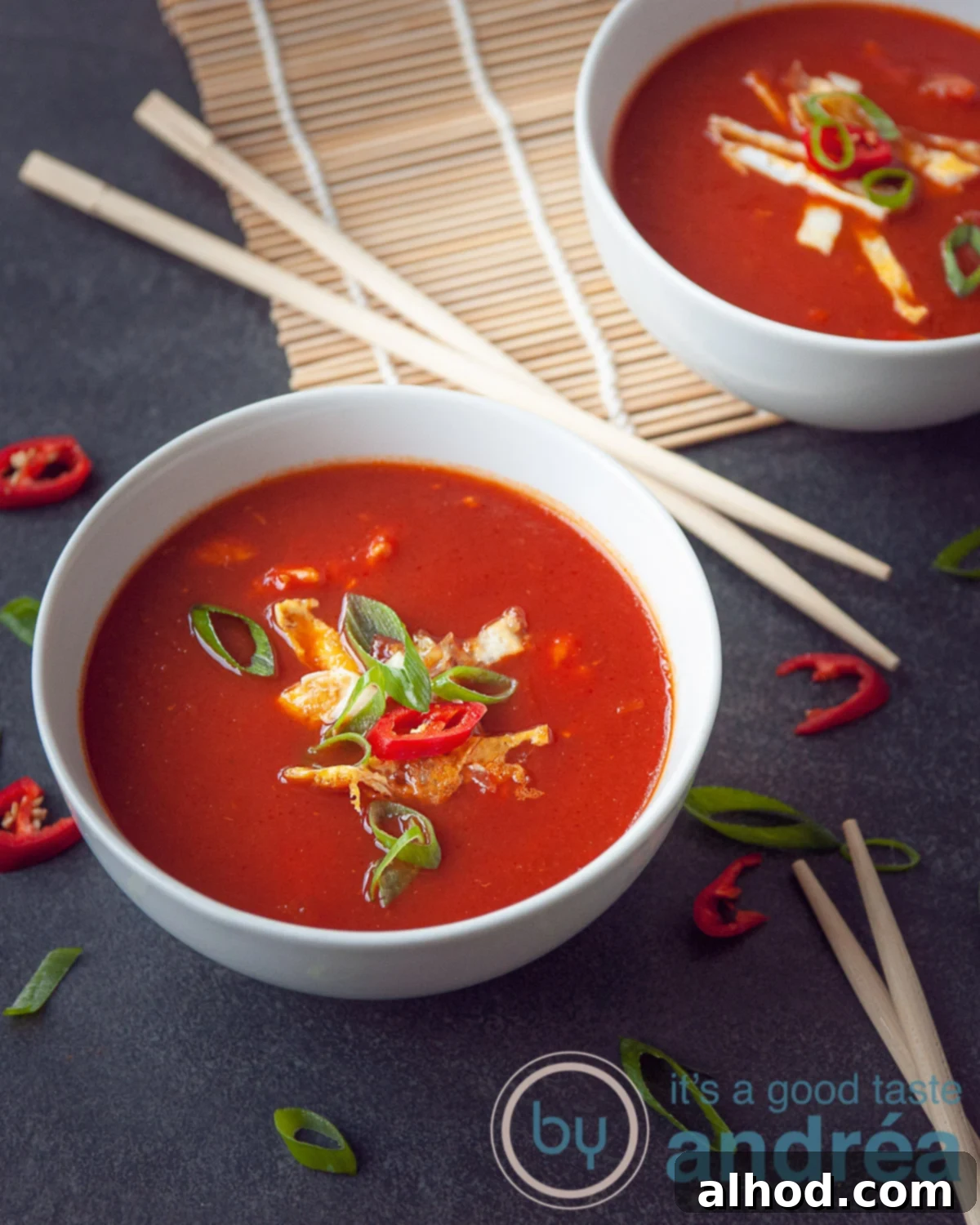 7 Incredible Asian Soups You Need to Taste 5 A grey background with two bowls filled with Asian tomatosoup with egg, spring onions and pepper. Some chopsticks and veggies scattered around