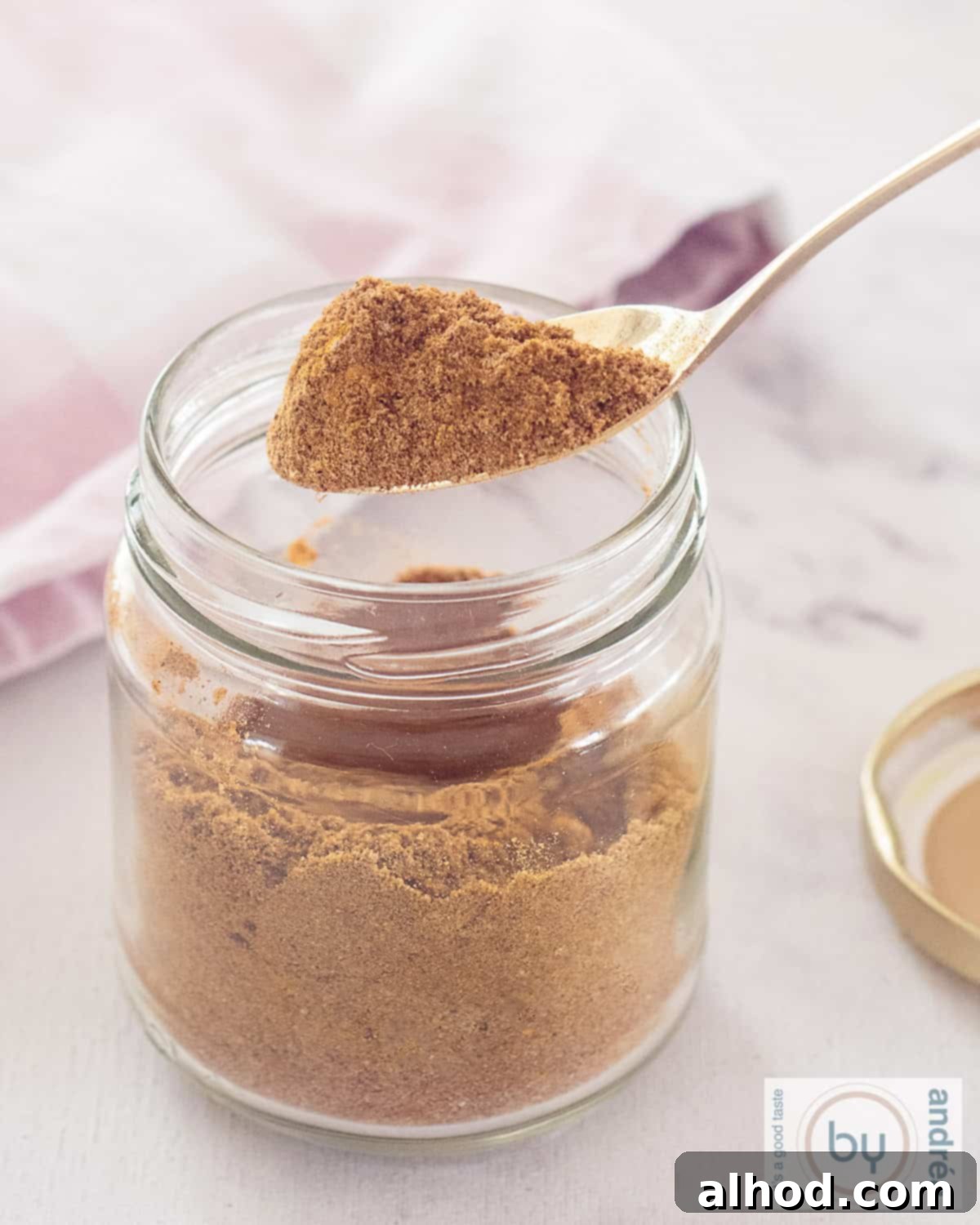 Authentic Homemade Madras Curry Blend 2 A tall photo featuring a glass jar filled with a rich, reddish-brown Curry Madras spice mix. A golden scoop, removed from the jar, holds a small pile of the aromatic spices, ready to be used.