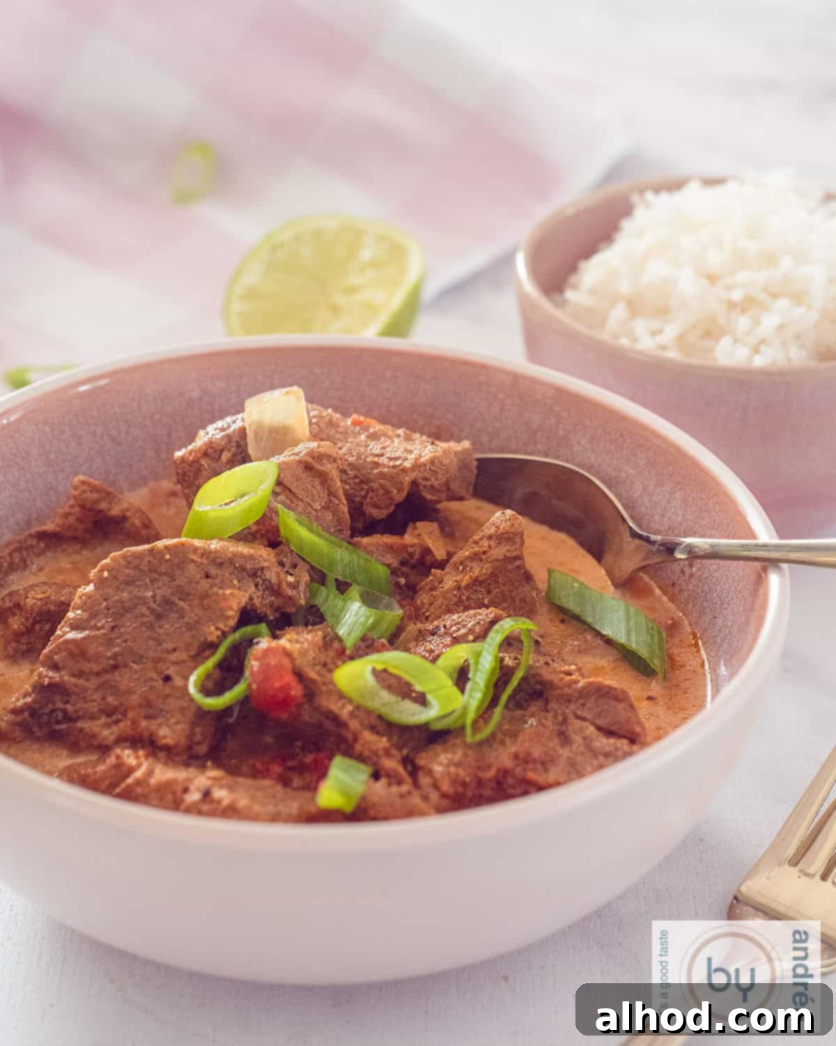 Tender Slow Cooker Beef Madras Curry 2 A vibrant pink bowl of slow-cooked beef madras curry, generously garnished with fresh spring onions. In the soft-focus background, a complementary bowl of steaming white rice awaits.