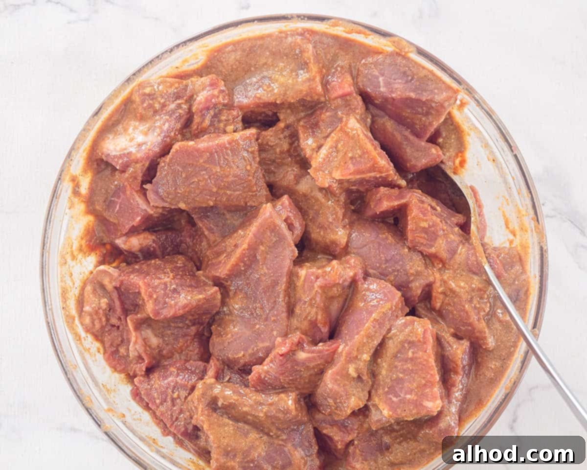 Tender Slow Cooker Beef Madras Curry 6 A transparent glass bowl holds chunks of beef generously coated in a rich, orange-hued marinade, ready for cooking.