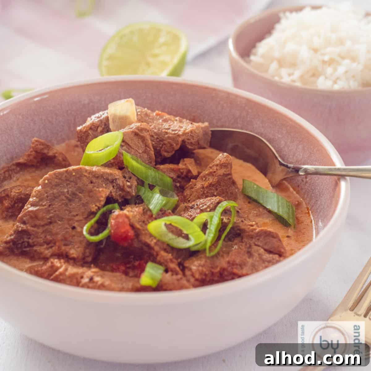 Tender Slow Cooker Beef Madras Curry 10 A square bowl filled with curry madras and spring onion. A pink bowl filled with rice diagonally behind it.