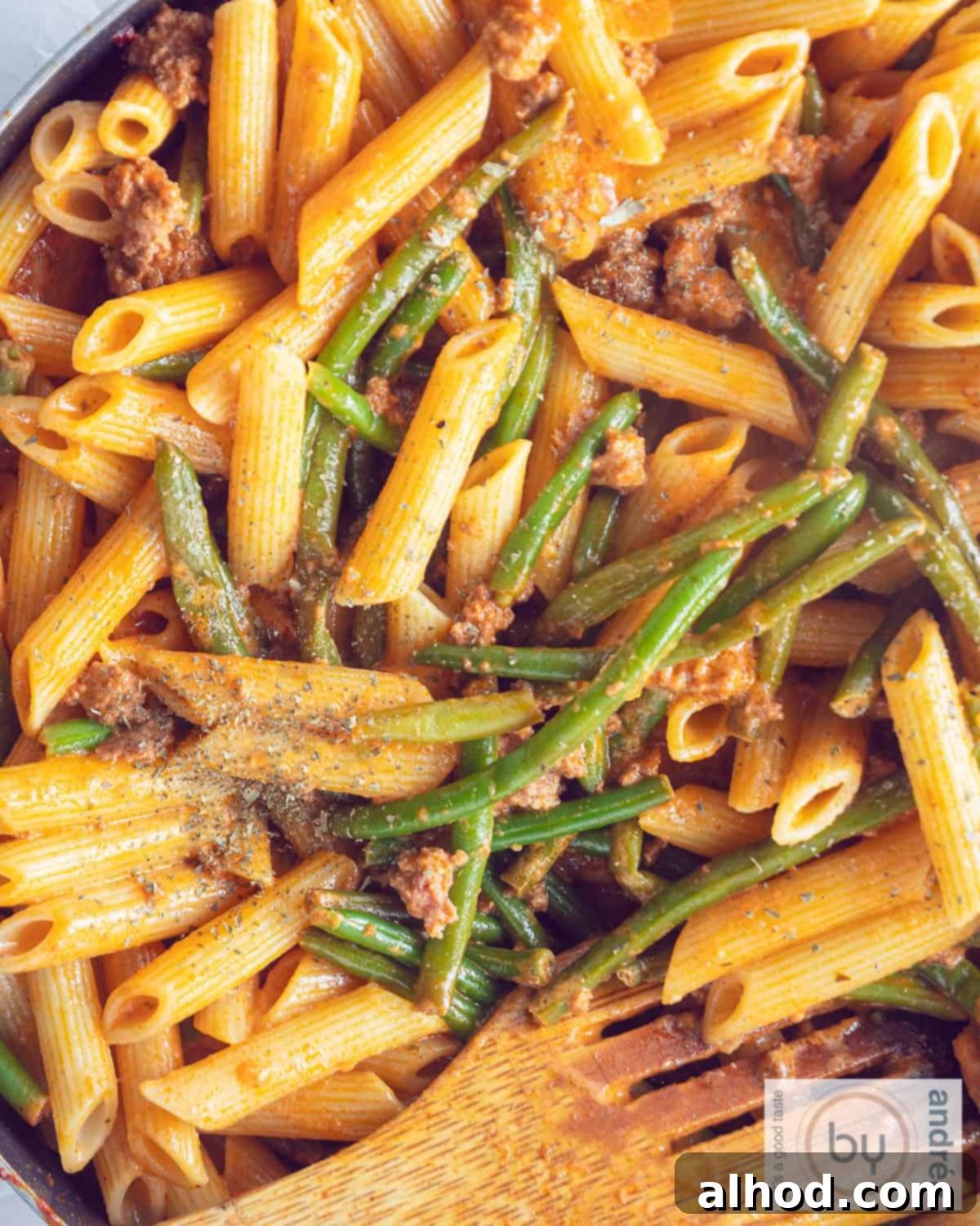 An elevation photo of a pan filled with penne alla vodka, minced meat and green beans. A wooden spoon is in the pan.