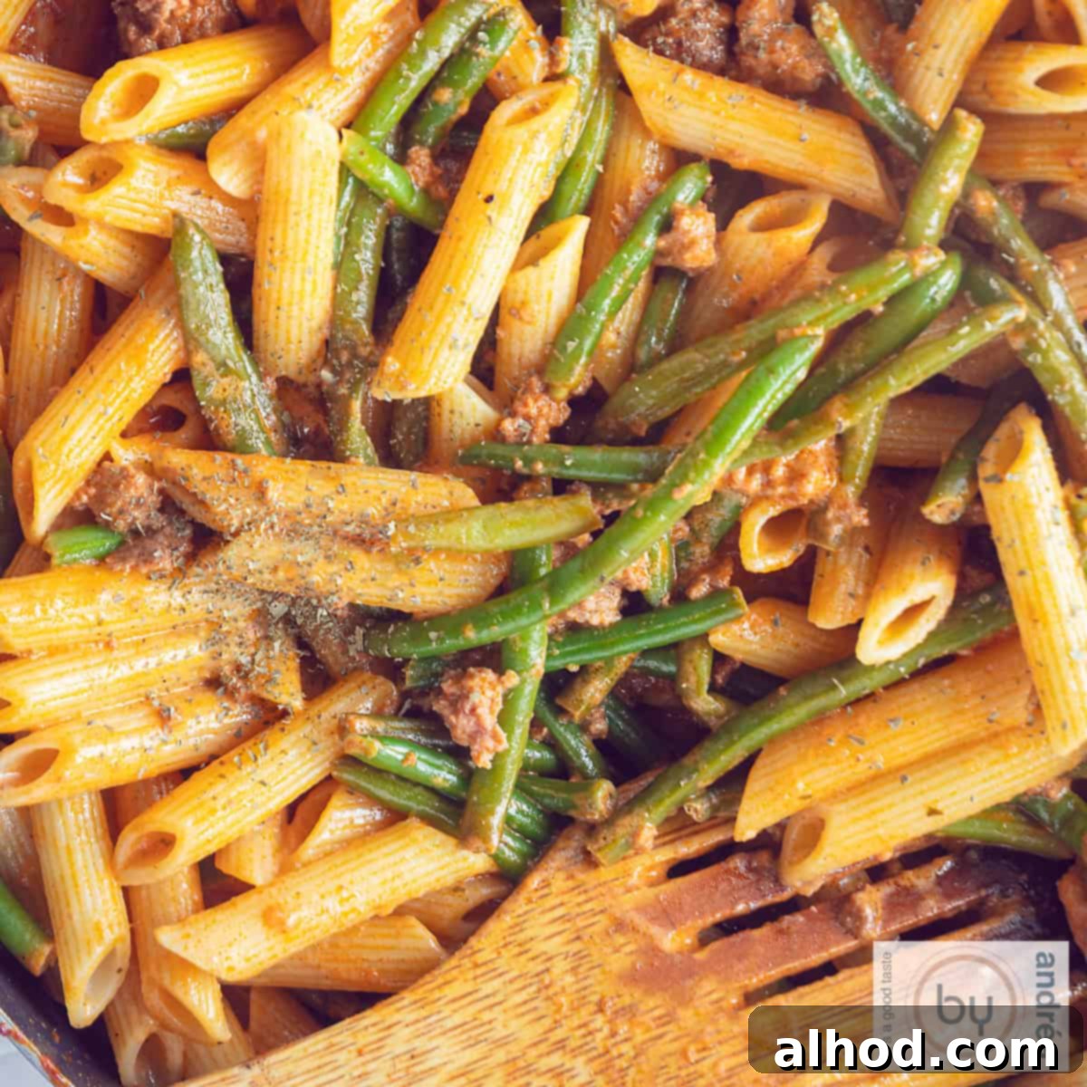 A square photo of a pan filled with penne alla vodka, with green beans and minced meat. A wooden spoon is in the pan.