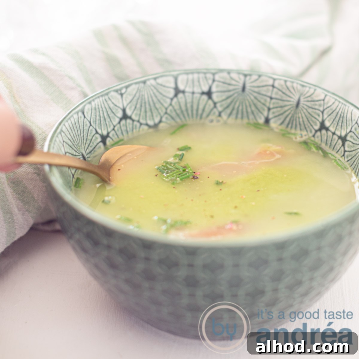 9 Delightfully Light Soups Packed with Fresh Vegetables 3 A square photo with a green bowl filled with green pea soup, Serrano ham, cream and chives. A hand takes a bite. A white background.