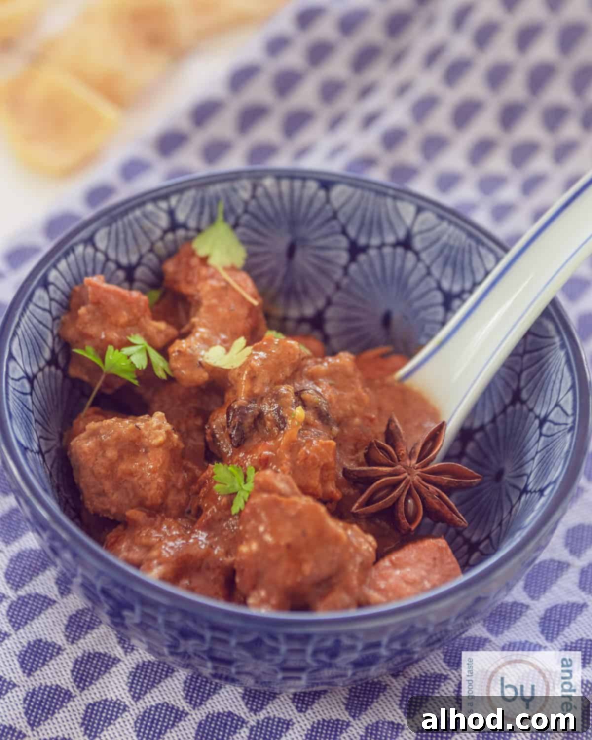 A blue bowl on a blue and white background filled with Indonesian rendang with a garnish of star anise and parsley
